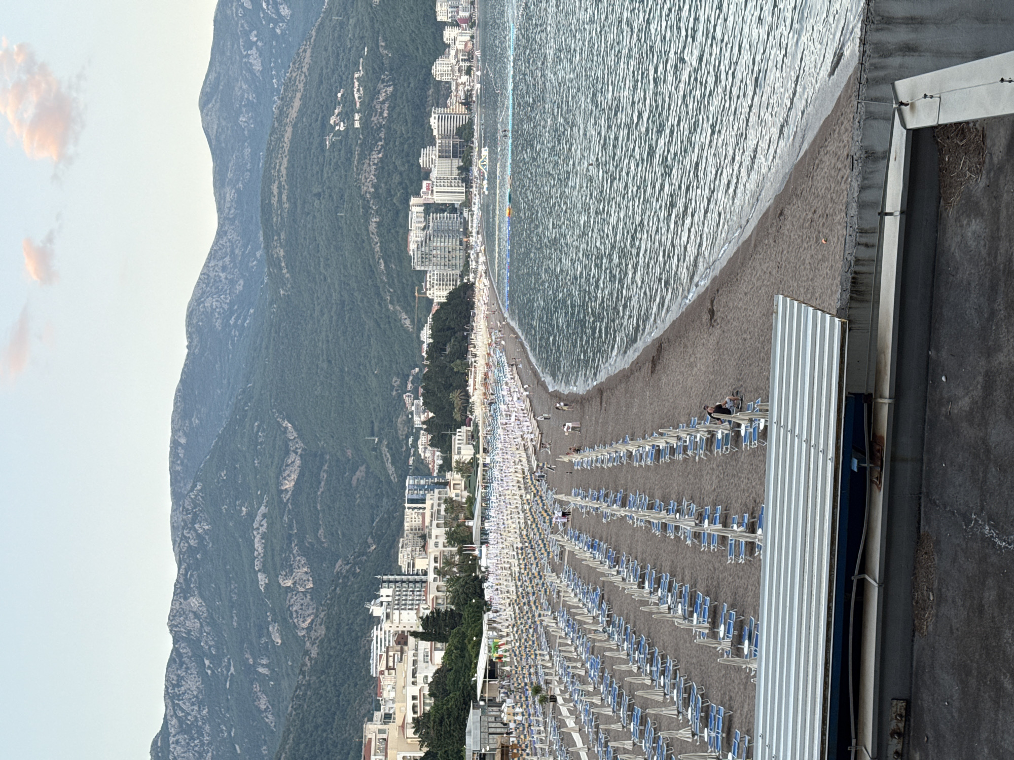 The image displays a coastal scene at the city of Boreti, Montenegro, featuring a densely packed beach with numerous rows of lounge chairs and umbrellas. The beach is situated along a body of water, likely the Adriatic Sea, which exhibits moderate waves and a shimmering surface reflecting the sky. In the background, a range of hills and mountains rises, densely covered in green vegetation. A line of buildings, indicative of a resort town, runs along the coastline between the beach and the mountains. Several people are visible on the beach, either walking or seated, and a few are in the water. A floating structure, possibly a water park element, is present in the sea. The lighting suggests it is either early morning or late afternoon, with soft, diffused light and a pale sky with a few pinkish clouds. In the foreground, a portion of a structure with a corrugated metal roof is visible, suggesting the vantage point is from a higher elevation or an adjacent building.