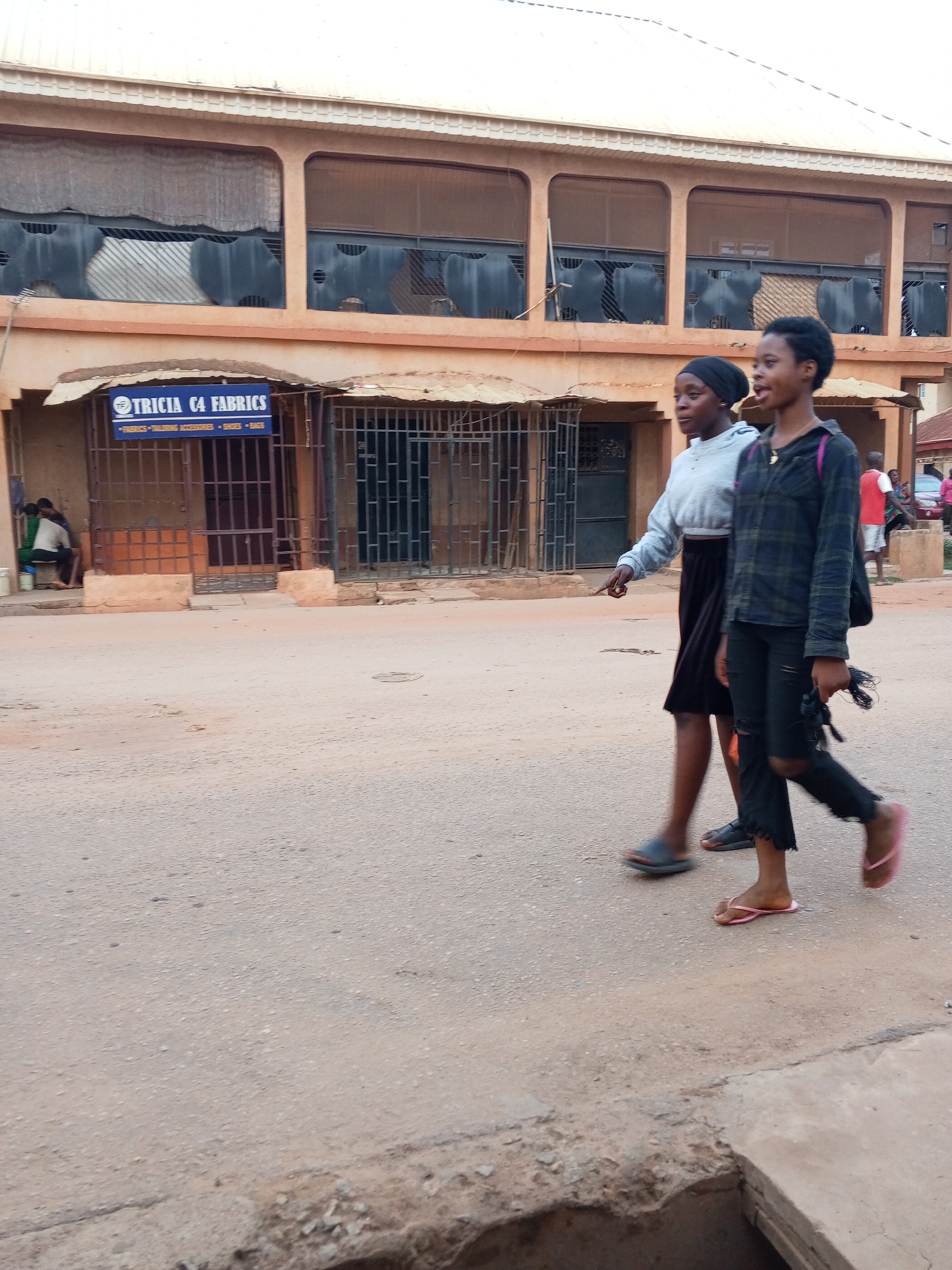 Two young women walk along a street in Sabon Tasha, Nigeria. The woman on the left wears a black hijab, a light gray long-sleeved shirt, and a black skirt. She points with her left hand. The woman on the right wears a dark plaid button-down shirt, torn black jeans, and pink sandals. She carries a black bag over her shoulder and an object in her right hand. They walk on a wide, dusty road. Behind them is a two-story building with bars on the windows on the first floor. A sign reading "TRICIA C4 FABRICS" is visible. People sit outside a building in the background on the left, and others stand on the right.