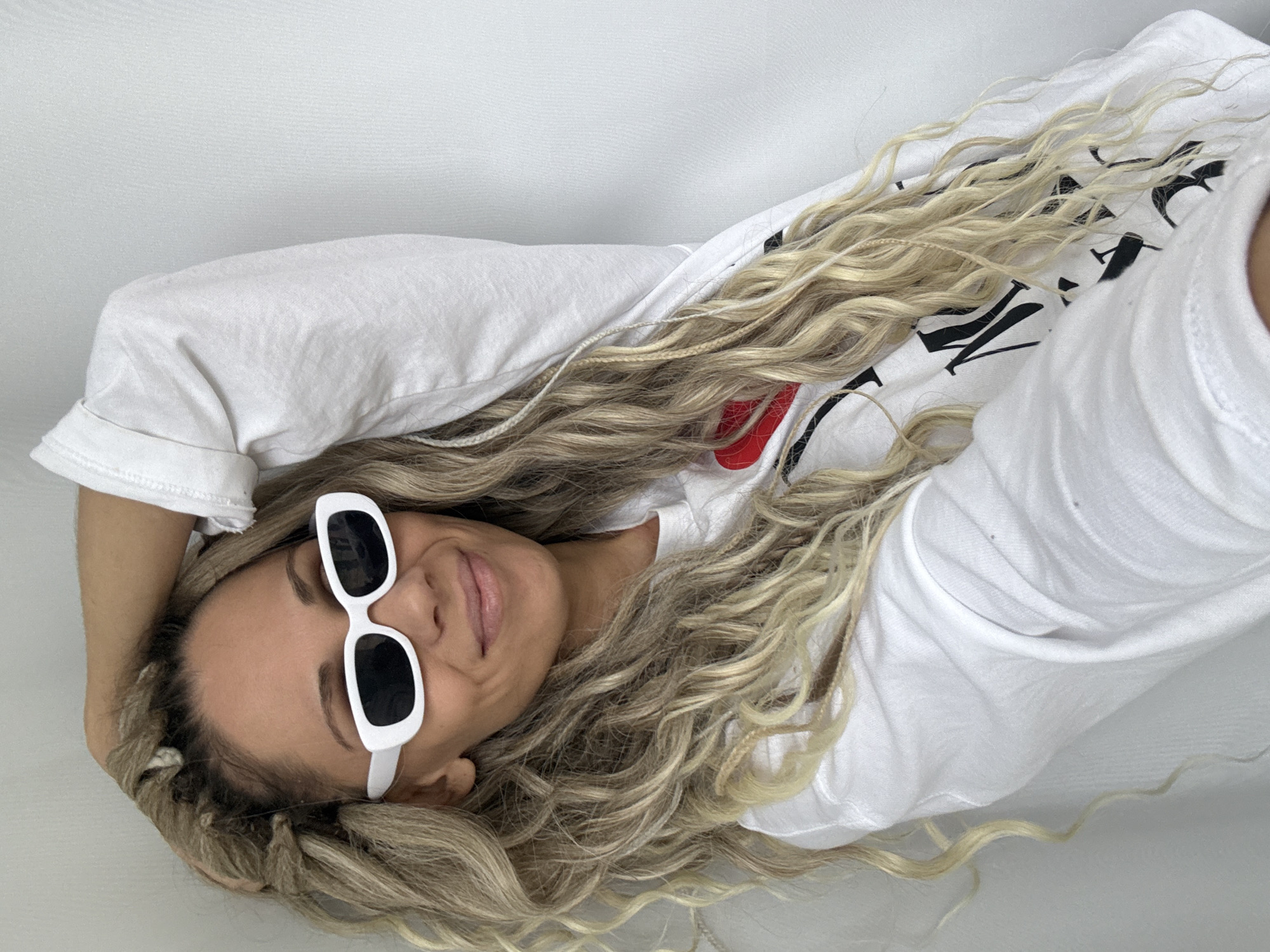 A woman with blonde hair and white sunglasses is lying on a white surface. She is wearing a white t-shirt with black lettering and a red heart design. Her head is resting on her hand. The location is not definitively identified as Rusanovo, Russia, based on the visual information in the image.