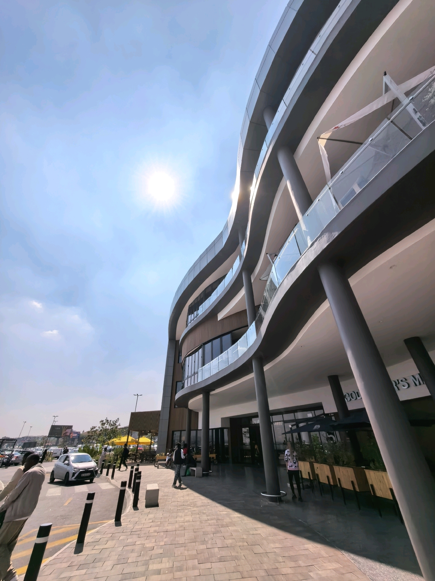 In Randburg, South Africa, on a bright, sunny day with a clear blue sky, the exterior of a modern, multi-story building is captured. The building features a curved facade with dark gray, wavy horizontal elements and glass balconies on multiple levels. Large, cylindrical gray columns support the upper floors, and the ground floor has large glass windows and doors, suggesting retail or dining establishments. A sign above the entrance reads "COLES M".
The foreground shows a paved pedestrian walkway with a textured brick pattern. Several black bollards and a concrete block are lined up along the edge of the walkway. To the left, a street with yellow lane markings is visible, where a white car is driving. More cars are parked in the background in a lot. Several individuals are present: two people are walking on the sidewalk, and another person is standing near the entrance of the building. Further back, near yellow umbrellas set up outside, more people are visible. The overall environment suggests a commercial or recreational area in a city.