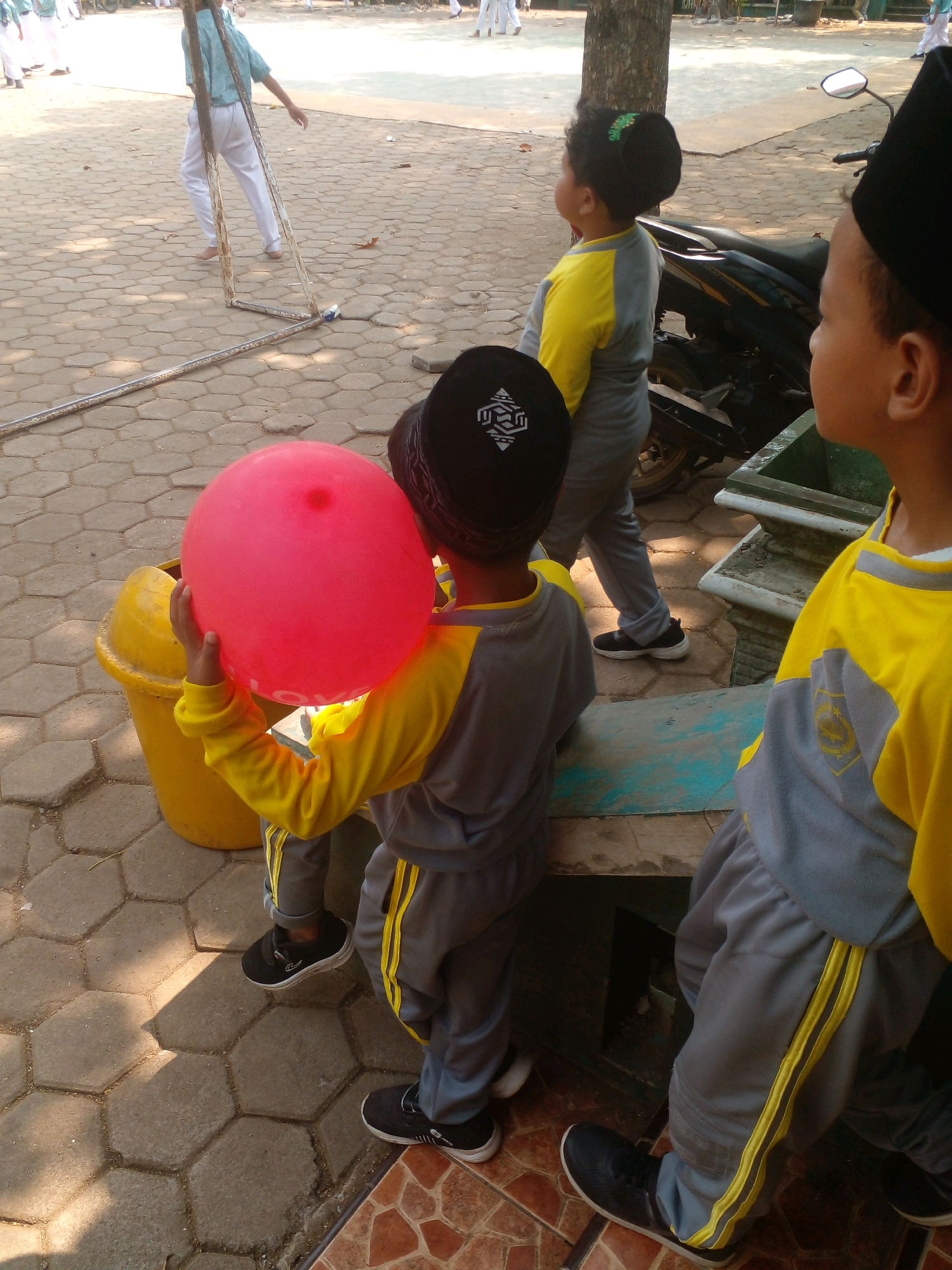 The image depicts a group of young boys in an outdoor setting, likely a schoolyard in Banyuputih, Indonesia, given the surrounding elements. In the foreground, a boy wearing a black cap and a yellow and gray uniform is holding a large pink balloon. To his right, another boy, also in a similar uniform, is partially visible from the side, facing away from the camera. Behind them, a scooter is parked, and in the background, a person in a light blue shirt and white pants is walking, possibly on stilts or a similar apparatus. The ground is paved with hexagonal tiles, and a yellow trash can is situated near the boy holding the balloon. The overall scene suggests a casual outdoor gathering or activity.