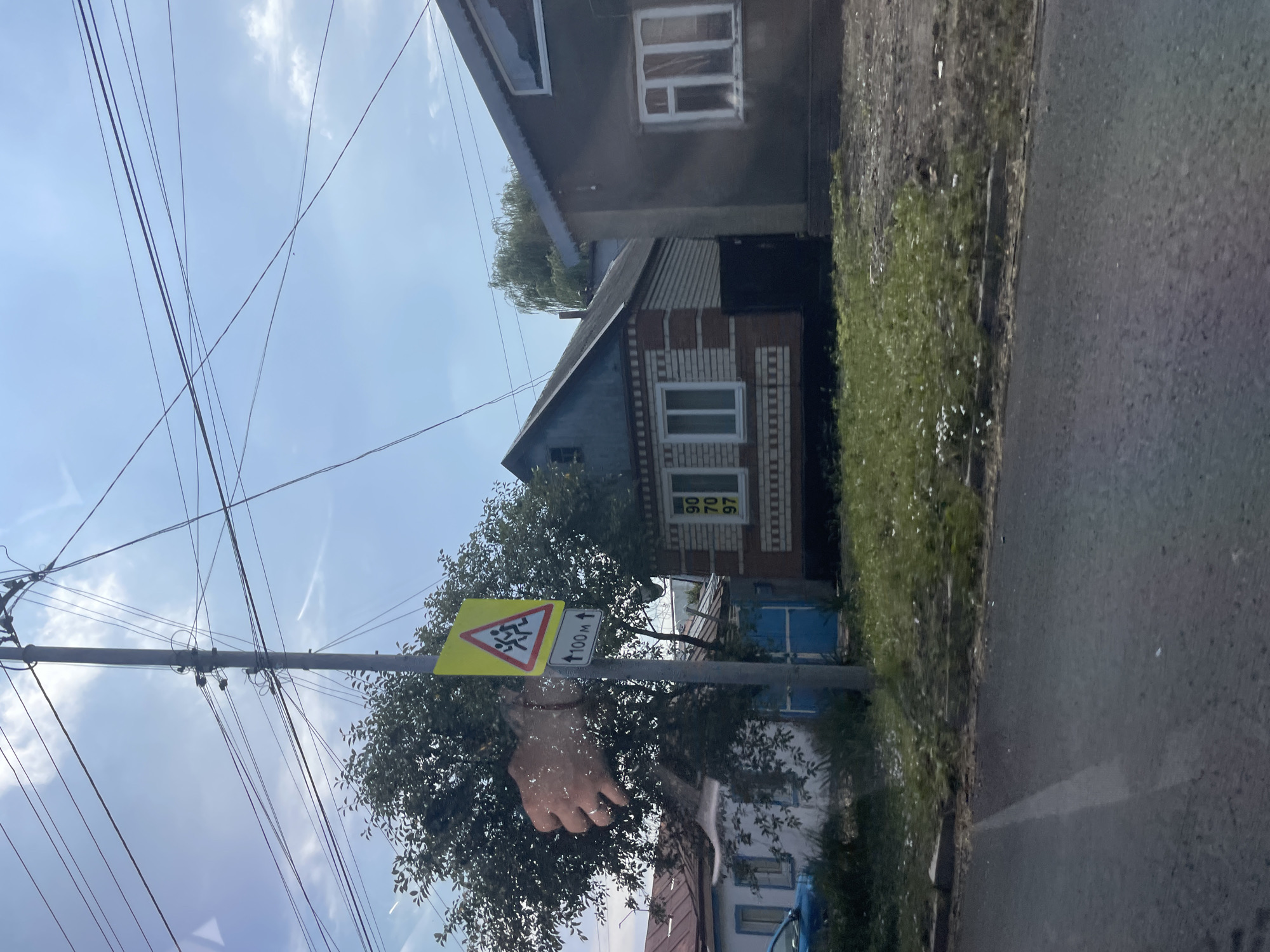 A roadside view shows a street in Stavropol, Russia, with houses lining the right side of the road. A utility pole with multiple wires stands on the left, adorned with a yellow triangular warning sign depicting children crossing and an additional sign indicating "100 m" with an upward arrow. The houses are single-story structures with tiled roofs, one with red brick accents. A blue fence is visible behind the utility pole. The foreground features a paved road with a grassy verge, dotted with small white flowers. The sky is partly cloudy.