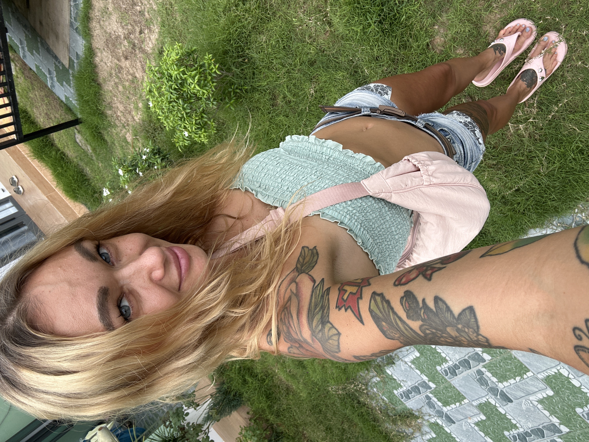 This media file is a high-angle selfie of a woman taken outdoors in a sunny environment, consistent with the provided location context of Mũi Né, Vietnam. The woman, who is the central subject, has long, flowing blonde hair and bright blue eyes, looking directly at the camera with a subtle, relaxed smile.
She is dressed in a light green, ruffled crop top that exposes her midriff, and light-wash denim shorts cinched with a brown belt. A light pink fanny pack or crossbody bag is worn across her chest. Her arms and legs are visibly covered with tattoos featuring various designs, including elaborate floral or leafy patterns on her arms. On her left foot, a small elephant design is visible beneath the strap of her light pink flip-flop.
The background consists of a well-maintained green lawn with some areas of patchy grass, alongside small, lush green bushes adorned with delicate white flowers. A distinctive paved path or patio area, characterized by a geometric pattern of grey and white tiles, is visible in the lower right and upper left portions of the image. Hints of a building, possibly with a dark frame, can be seen in the background, suggesting a landscaped property or resort setting. The bright lighting indicates it is daytime. There is no visible text in the image.