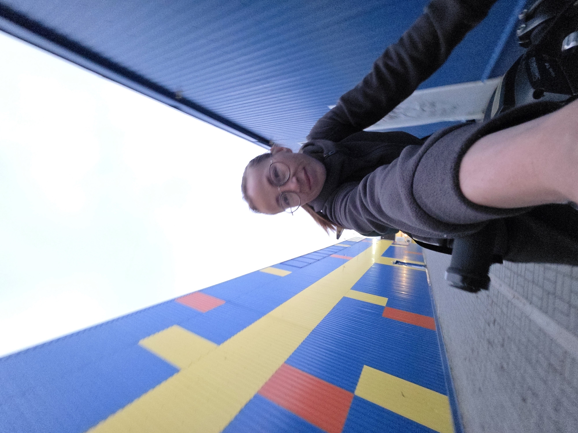 The image captures a selfie of a woman in the city of Chaikovsky, Russia, taken from a low angle with a wide-angle lens. The woman is positioned slightly right of center, looking directly at the camera with a faint smile. She has light skin, light-colored hair pulled back, and wears round eyeglasses along with a dark grey, high-collared, zip-up fleece jacket. Her right arm is extended, holding the camera, with parts of her arm visible in the lower center. In the lower right foreground, components of a bicycle are visible, including a black handlebar grip and a shifter mechanism bearing the "SHIMANO" brand. The woman appears to be situated on a paved pathway made of light grey interlocking pavers, which recedes into the background between two large structures. The building on the left features a corrugated blue metal facade decorated with a large, repeating geometric pattern of yellow and red rectangular shapes. The building on the right also presents a corrugated blue metal facade, appearing uniform in color. The sky above the buildings is a bright, uniform white.