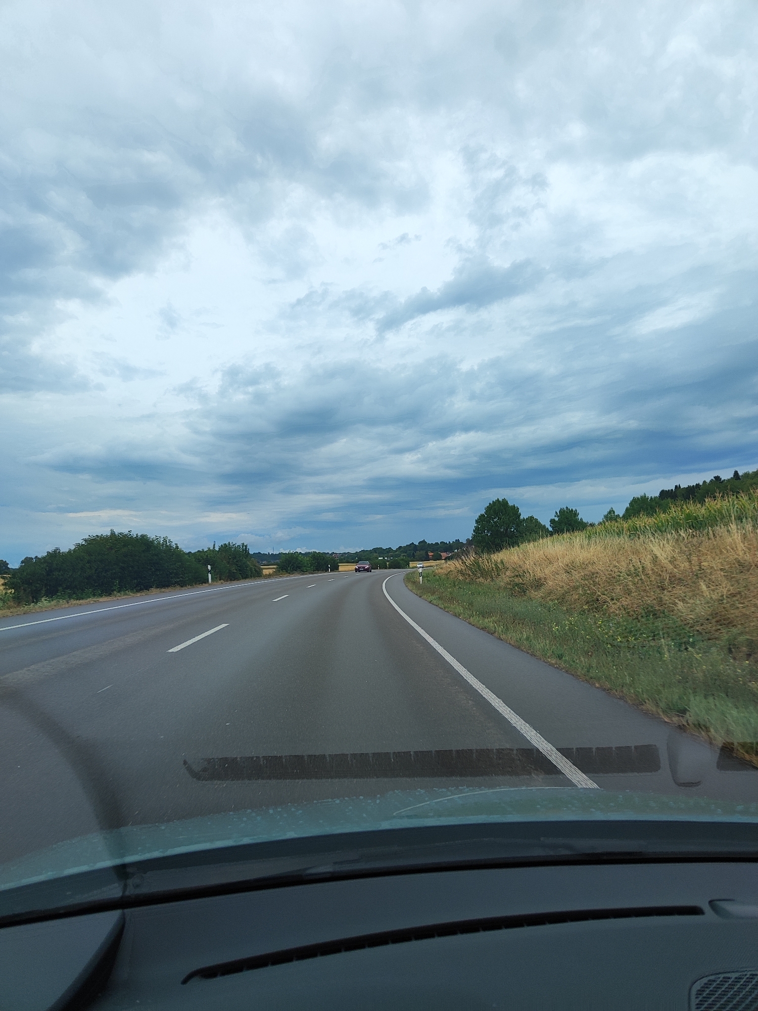 The image shows a view from inside a vehicle, looking out onto a two-lane road that curves to the right. The road surface is asphalt and appears wet, with white lane markings. To the left of the road, there are green bushes and trees. To the right, there is a field of dry grass and a cornfield, with more trees and some houses visible in the distance. The sky is overcast with dark, dramatic clouds. In the distance, on the right lane of the road, a red car is visible. The location is the B3 highway in Friesenheim, Germany.