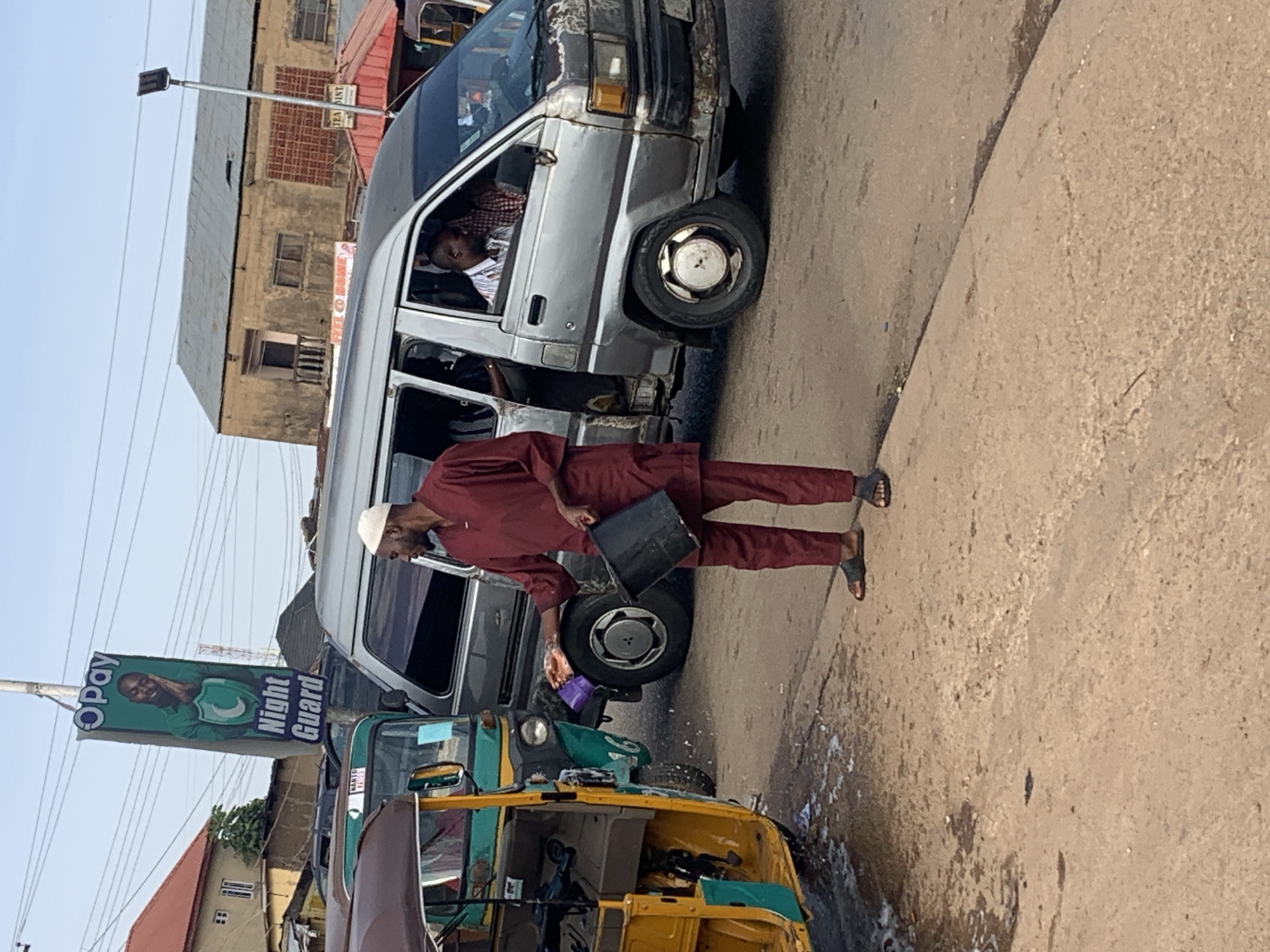 A medium shot captures a street scene in Gura Top, Nigeria. A man in a maroon tunic and pants, wearing a white cap and sandals, is in the foreground, holding a black bucket and what appears to be a purple cloth in his outstretched left hand. He is facing towards the left, seemingly interacting with a yellow and green tuk-tuk. White foam or suds are visible on the ground near the tuk-tuk's wheel, suggesting a washing activity. Behind the man, a silver minivan is parked. The driver, wearing a striped shirt, is visible through the open driver's side window. Another passenger is partially visible in the passenger seat. The background shows buildings with weathered facades and a pole with an OPay "Night Guard" advertisement. Power lines crisscross the sky. The road surface is paved, and the overall lighting suggests daytime.