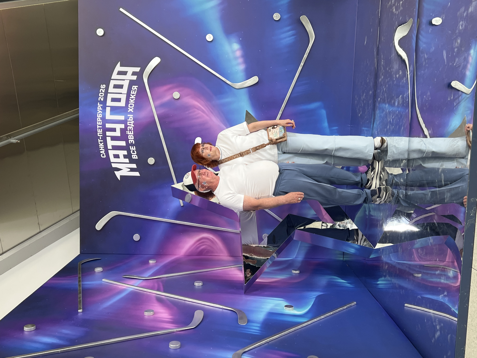The image depicts two individuals, a man and a woman, posing in front of a large, colorful backdrop featuring a cosmic-themed design with stylized hockey sticks. The backdrop is predominantly blue and purple, with streaks of pink and white, creating an aurora borealis effect. The text on the backdrop reads "САНКТ-ПЕТЕРБУРГ 2026" (Saint Petersburg 2026) and "МАТЧ ГОДА" (Match of the Year) along with "ВСЕ ЗВЁЗДЫ ХОККЕЯ" (All Stars of Hockey).
The man, on the left, is wearing a white t-shirt, blue pants, and a baseball cap adorned with red and white stars. He has a ring on his left hand. The woman, on the right, is wearing a white t-shirt, light blue pants, and a white cap. She has a crossbody bag with a colorful pattern. Both individuals have small star-shaped stickers on their faces.
The foreground consists of a reflective, mirrored surface that captures distorted reflections of the individuals and the backdrop. This mirrored surface is part of an exhibit or installation, possibly in an indoor public space. The overall impression is that of a promotional event or an interactive photo opportunity related to the "All Stars of Hockey" event scheduled for Saint Petersburg in 2026. The environment appears to be a well-lit indoor setting, likely a venue or exhibition hall.