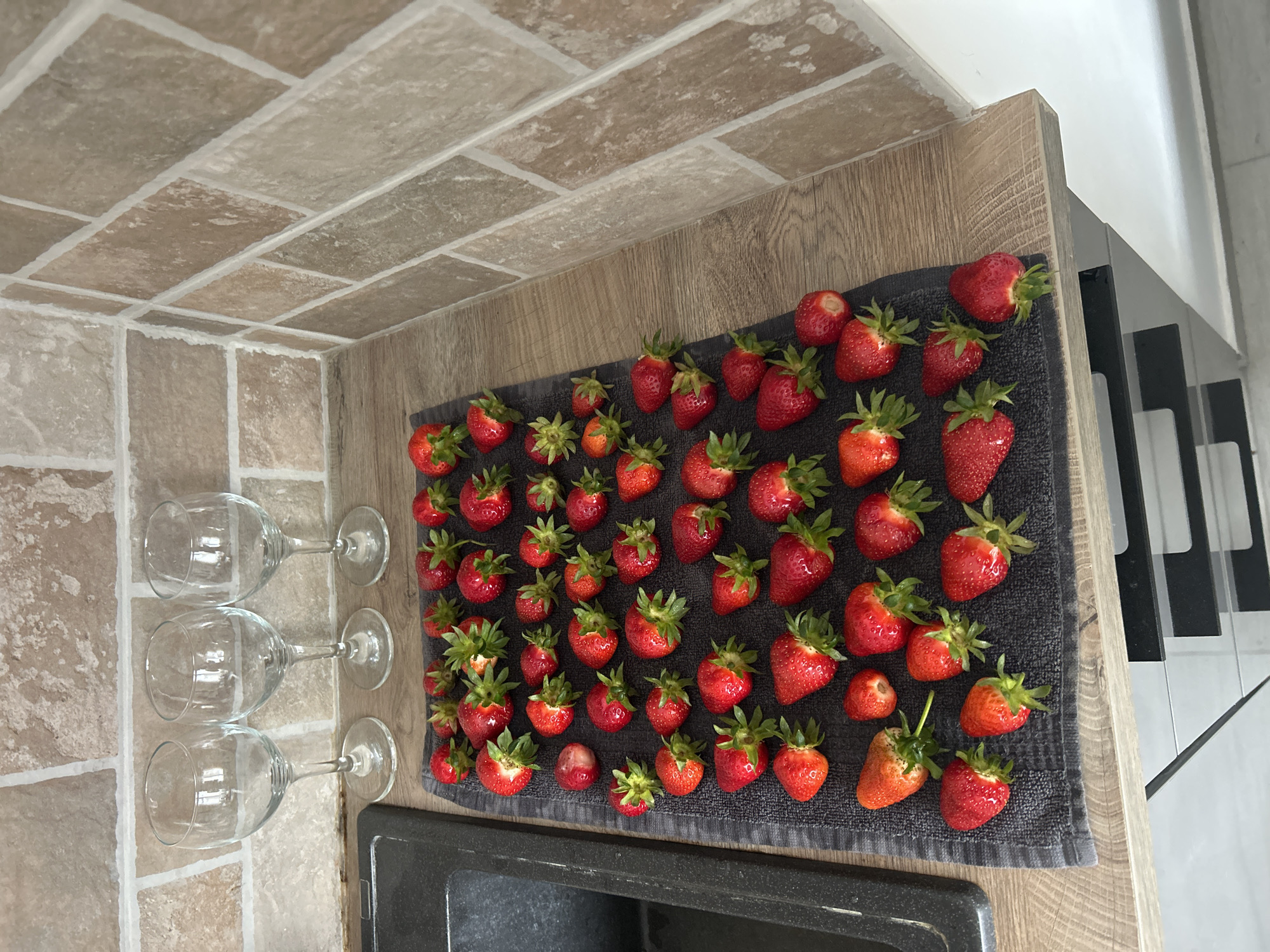 The image depicts a countertop arrangement. A grey textured towel displays approximately 50 fresh strawberries with green stems and calyxes. Adjacent to the towel are three empty wine glasses. The countertop has a light-brown wood-grain finish. The background includes a brick-patterned wall and a partial view of a black and white object, possibly a keyboard.
The location is inferred to be Budva, Montenegro, based on the presence of grapes, which are grown in that region of the world.