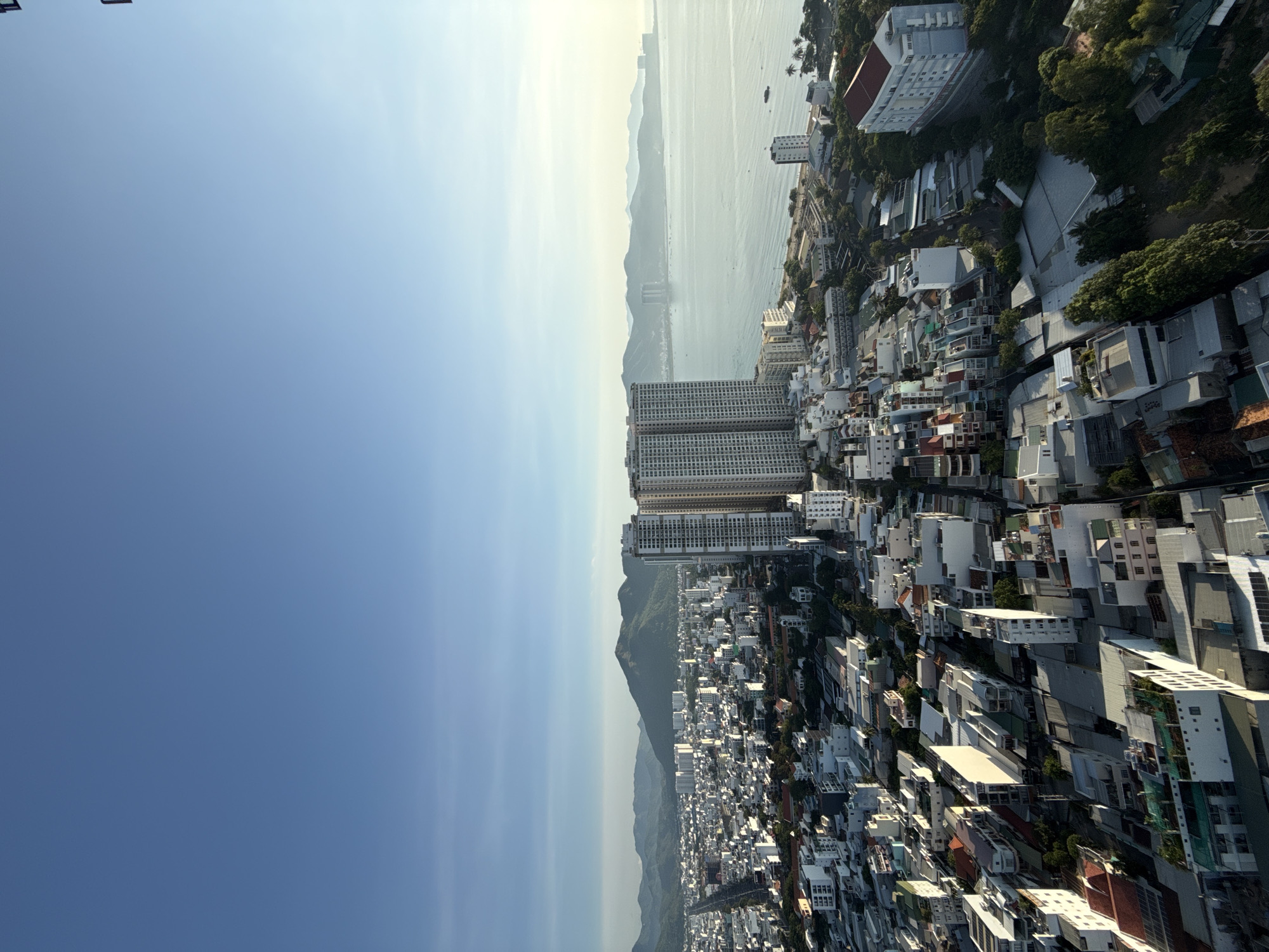This image is an aerial view of a coastal city in Nha Trang, Vietnam. In the foreground, a dense urban landscape of buildings of varying heights, primarily white and gray, stretches across the frame. Many of these buildings appear to be residential or commercial, with flat roofs and balconies. Some buildings have red-tiled roofs. Further back, a wide, tree-lined road is visible, leading towards the coast. Dominating the mid-ground are two tall, modern apartment buildings, their facades featuring many windows. Beyond these structures lies the Nha Trang Bay, a calm body of water dotted with a few small boats. In the distance, a hazy mountain range forms the horizon, with the sky above a clear, pale blue, transitioning to a lighter hue near the horizon. The overall scene depicts a sunny day with clear visibility.