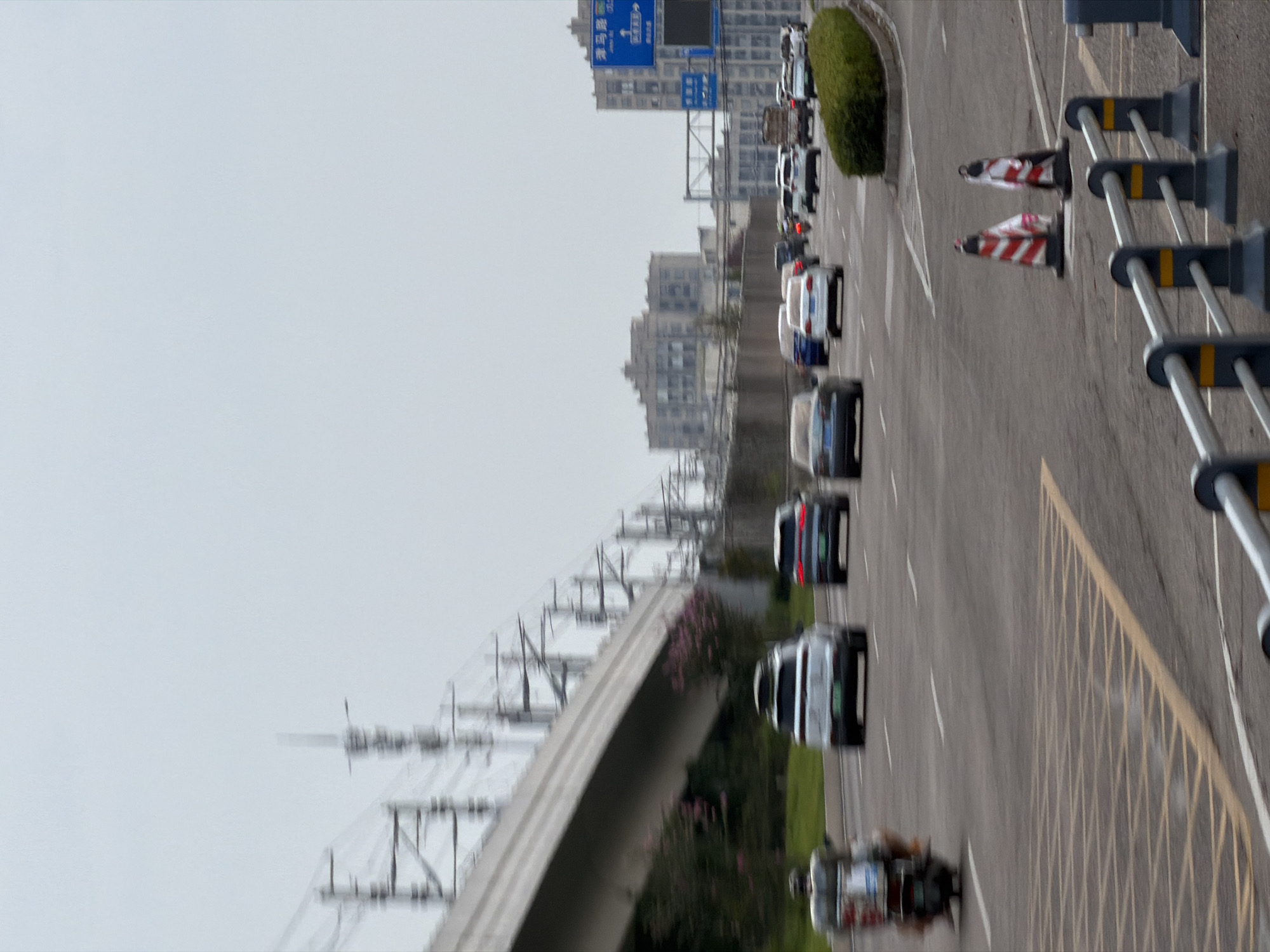 The image shows a street scene in Jiangjin, China, on an overcast day. A multi-lane road is depicted with several vehicles in motion. On the left, a scooter carrying a person is moving in the foreground. Further down the road, a white SUV with a roof rack is visible, followed by a dark-colored SUV. Other cars and trucks are scattered across the lanes. In the background, buildings and elevated train tracks with associated infrastructure are present. Traffic cones and barriers line the side of the road, suggesting ongoing roadwork or lane closures. The sky is a uniform light gray.