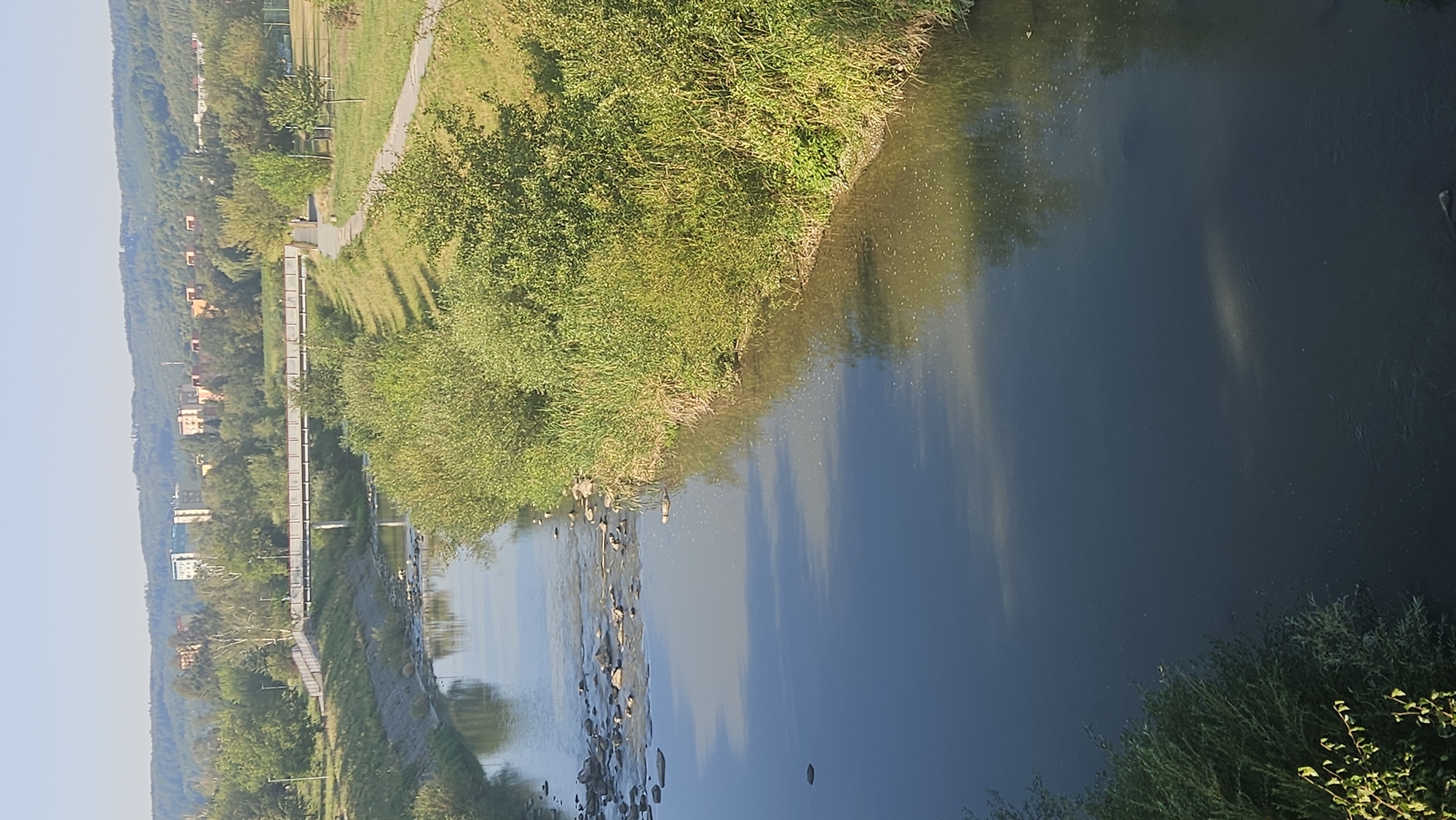 The image depicts a sunny, clear day in Prešov, Slovakia, featuring a river flowing through an urban park area. A pedestrian bridge spans the river in the mid-ground, with several individuals visible walking across it. The riverbanks are lined with lush green vegetation, including trees and bushes. In the background, a cluster of residential buildings is visible, set against a backdrop of rolling, tree-covered hills. The water in the river appears calm, reflecting the sky and surrounding greenery. The scene suggests a recreational area within the city, offering a natural escape amidst urban life.