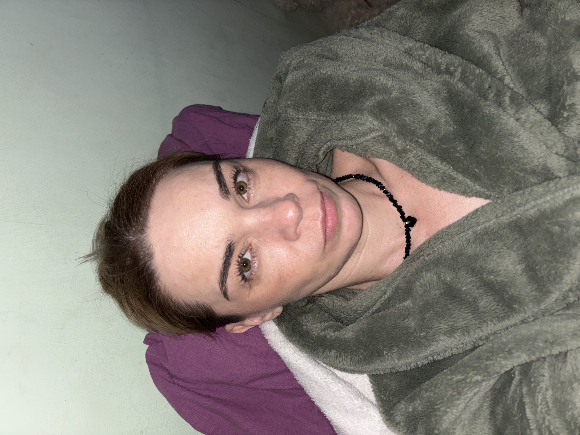 An eye-level indoor shot shows a woman lying down, looking directly at the camera with a calm expression. She has brown hair pulled back from her face, hazel eyes, and fair skin, with a natural or minimal makeup look. Around her neck, she wears a black beaded necklace with a small, dark heart-shaped pendant.
She is dressed in a plush, dark olive green or grey robe or blanket, which covers her upper body and arms. Below her head is a purple pillow or blanket, and further down, a white, textured blanket or towel is visible. The background consists of a plain, light greenish-grey wall on the left side of the frame. In the upper right corner, some indistinct dark shapes suggest an unidentifiable part of the room or ceiling. The lighting is soft and even, indicating an indoor setting with no specific time of day. No text is visible in the image.