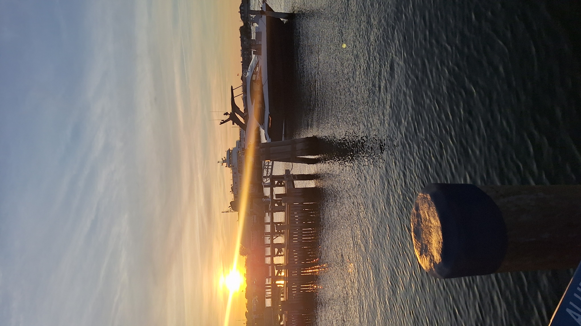 The image captures a serene sunset scene over a harbor in Nantucket, United States of America. The sun, a brilliant golden orb, is low on the horizon, slightly to the left of the center, casting a strong, warm glow across the water and sky.
The upper portion of the frame is dominated by the sky, which transitions from a soft, light blue at the top to vibrant orange and golden hues near the horizon. Wispy, elongated clouds are scattered across the sky, catching the sunset's light.
In the midground, several boats, appearing to be yachts or ferries, are docked or moored along a wooden pier structure that stretches horizontally. The boats are largely silhouetted against the bright backlight, their forms creating dark shapes on the water. Details like masts, cabins, and rigging are subtly visible. The pier's wooden pilings are also silhouetted, with some illuminated by the direct sunlight. The water reflects the golden light, creating shimmering ripples and streaks, particularly in the areas directly in front of the sun.
In the immediate foreground on the right, a stout wooden post with a dark, rounded cap stands prominently. Its top and right side are bathed in the golden light of the setting sun. At the very bottom right corner, a small section of a blue sign is visible, bearing partial white text that appears to read "ALLU".
The overall impression is one of tranquility and beauty, characteristic of the "golden hour" during sunset. The water is calm, and there are no people visibly engaged in activity, reinforcing the peaceful atmosphere.