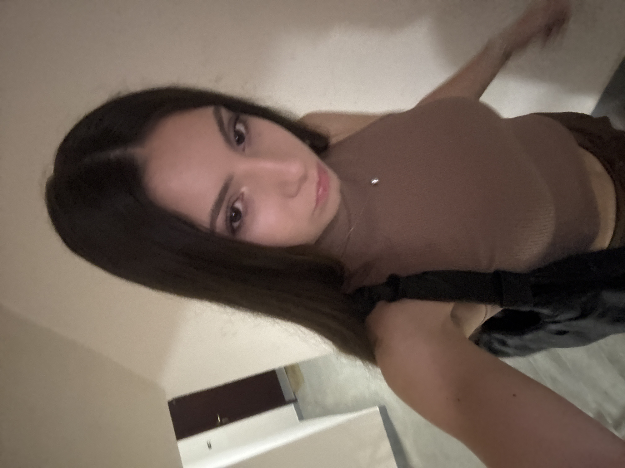 A young woman with long, dark hair is taking a selfie in a dimly lit indoor hallway, possibly in Varna, Bulgaria. She is wearing a sleeveless, ribbed, taupe-colored top and appears to be holding a black bag over her left shoulder. The hallway has plain, light-colored walls and a dark door is visible in the background on the left. The lighting is dim, casting a slight shadow on the woman's face.