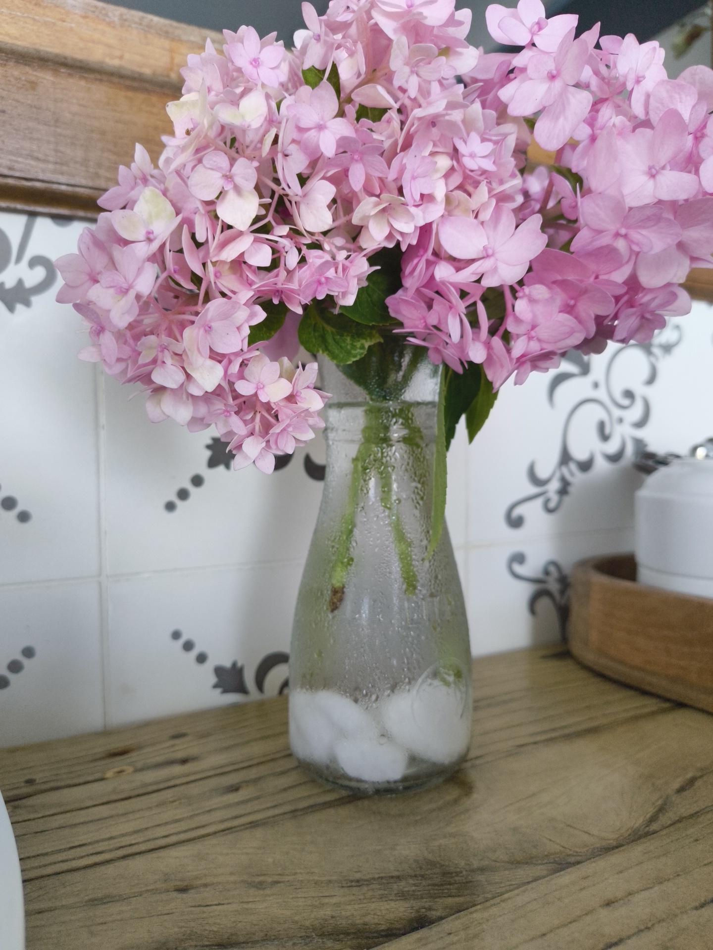 A close-up, eye-level shot captures a glass vase filled with water and ice, holding a bouquet of pink hydrangeas. The vase is placed on a wooden surface, and behind it, a tiled wall with a decorative pattern is visible. To the right, a wooden tray holding a white ceramic container is partially in view. The lighting is soft, creating a gentle atmosphere. The image does not provide any information about the city of Phu Thuy Ward, Vietnam.