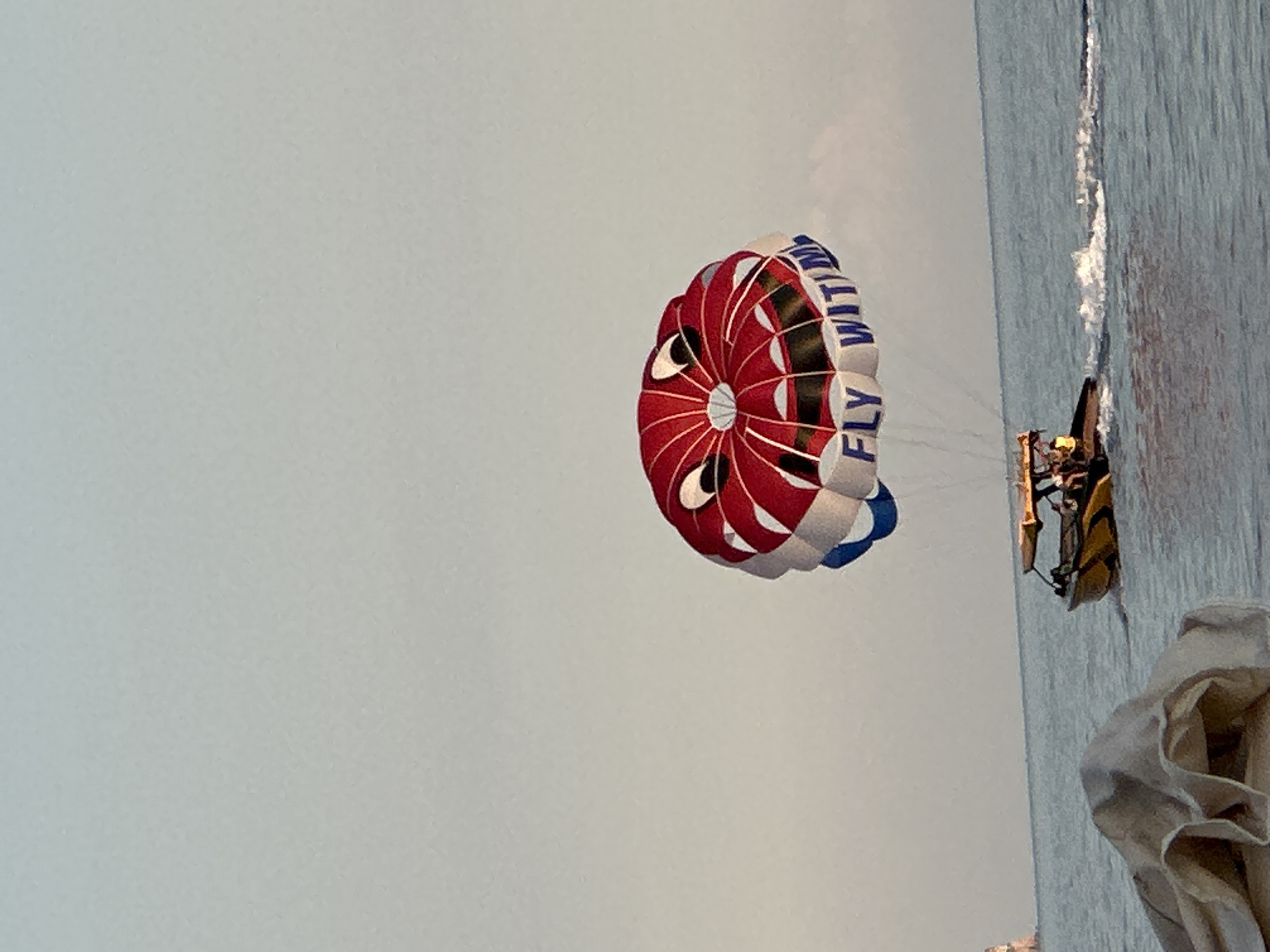 The image shows a parasail with a smiley face design with the words "FLY WITH ME" printed on it, suspended in the air over the Adriatic Sea. A yellow boat with a canopy is visible in the water, trailing the parasail and kicking up a wake. The sky is clear and pale blue, indicating daytime. The scene appears to be at a beach resort, possibly in Budva, Montenegro, given the presence of the sea and recreational water activities. In the foreground, a light-colored fabric is draped, obscuring part of the bottom left of the image.