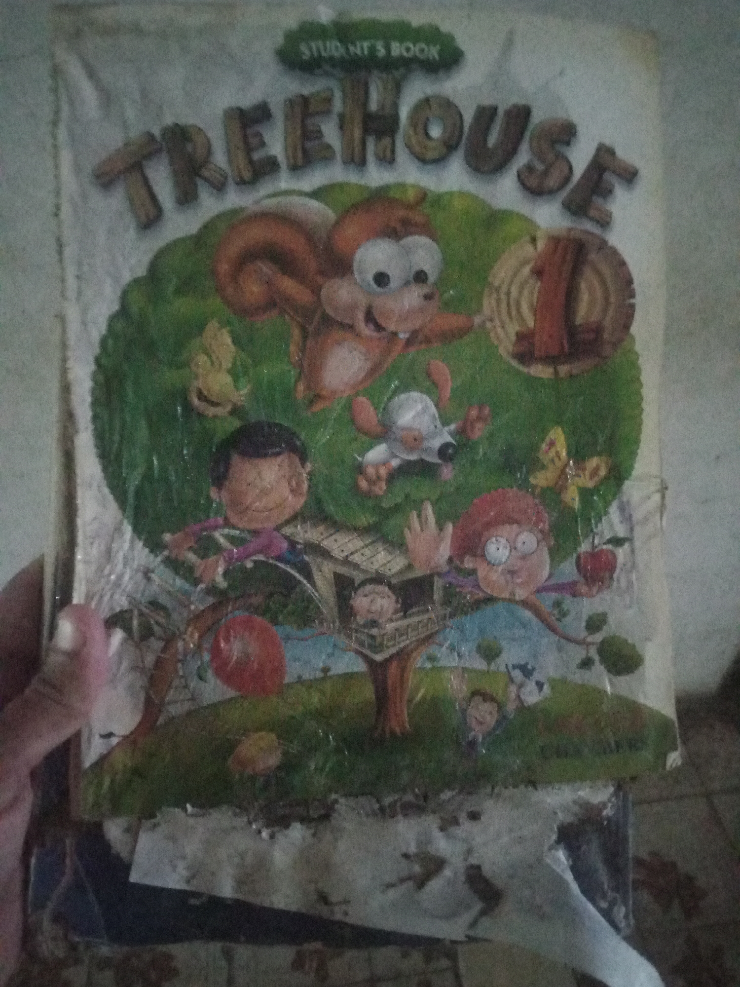 The image shows the cover of a children's book titled "TREEHOUSE 1". The cover features a colorful illustration with a cartoon squirrel at the top, a treehouse in the center, and various characters like children and a dog. The book is held by a person's hand, which is partially visible on the left side of the frame. The background appears to be a wall. The image is blurry and shows signs of wear and tear on the book cover, suggesting it is an old or frequently used item. The text "STUDENT'S BOOK" is also visible at the top of the cover. There is no information in the image that would allow for a determination of its location in Santa Ana, Venezuela.