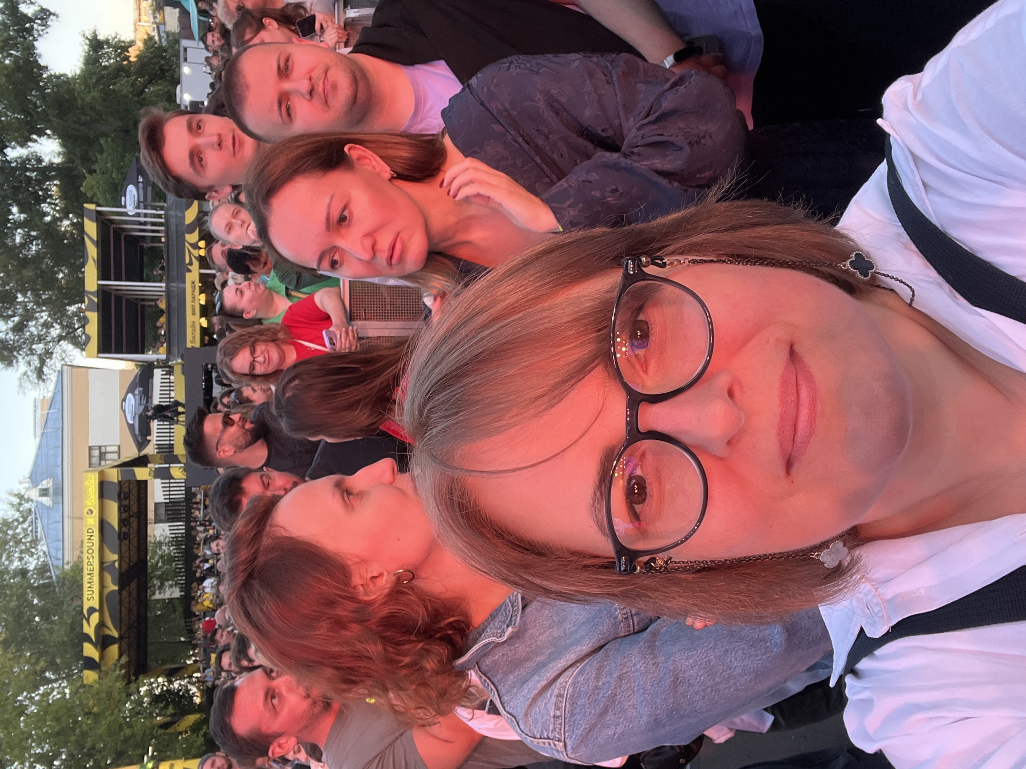 In Khimki, Russia, at a Summer Sound festival, a group of people are gathered, with the foreground dominated by a woman taking a selfie. She wears glasses with a decorative chain and a white shirt. Behind her, a crowd watches a stage, indicated by a yellow and black banner overhead that reads "SUMMERSOUND". The atmosphere appears to be that of an outdoor music event, with many individuals visible and a sense of shared enjoyment. The lighting suggests it is late afternoon or early evening.