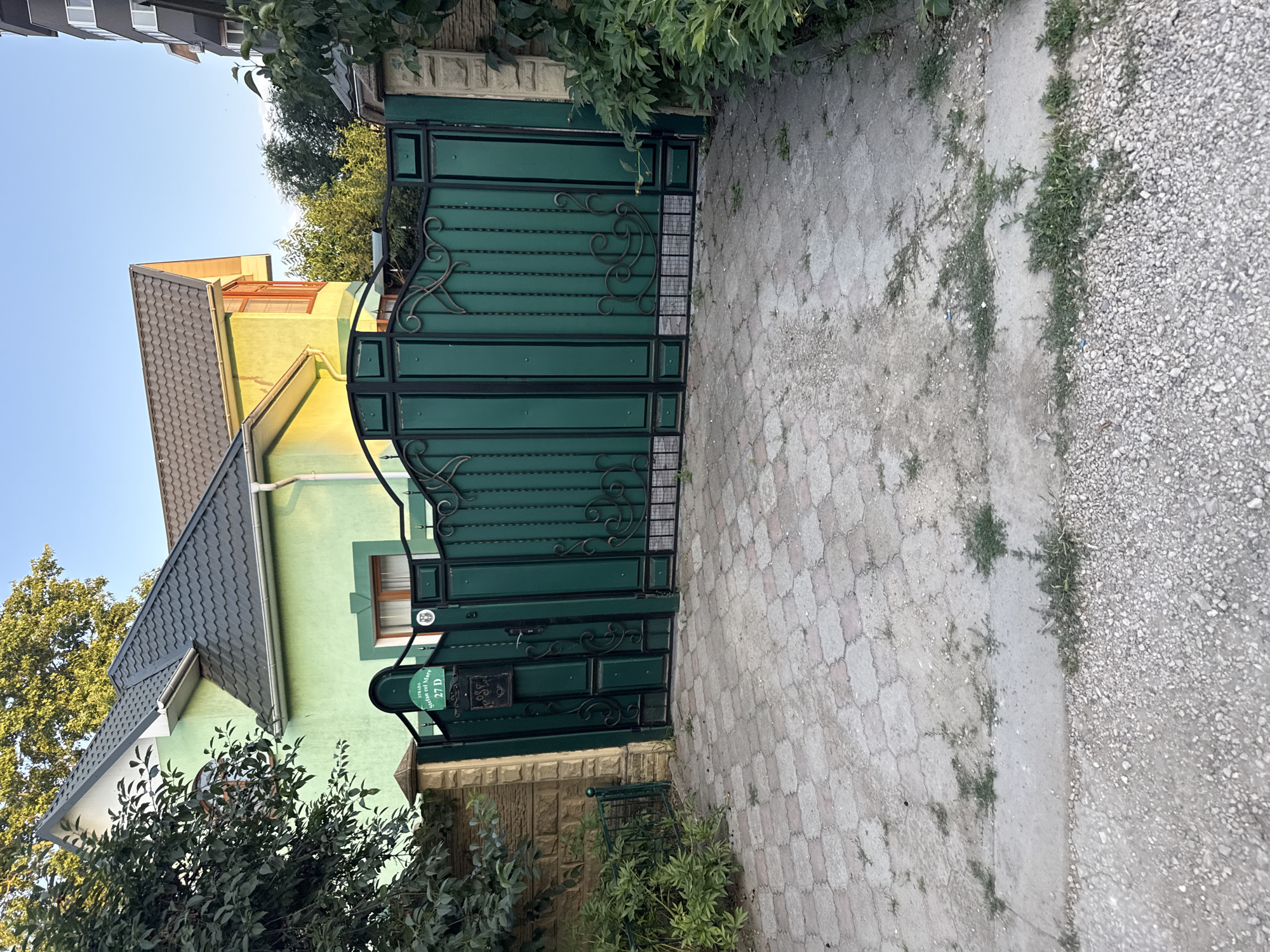The image depicts a green metal gate with decorative iron scrollwork, set against a backdrop of a light green house with a dark gray tiled roof. The gate appears to be closed. Adjacent to the gate is a pathway paved with interlocking stone tiles, bordered by a section of gravel. Green foliage from trees and bushes is visible, partially obscuring the house and framing the scene. A street sign with the text "STRADA MUNTENI NO 27 D" is attached to the gate. A mailbox is also mounted on the gate. The scene suggests an outdoor setting in a residential area, possibly during daylight hours with clear weather. The overall impression is one of a well-maintained, possibly suburban or rural, property entrance.