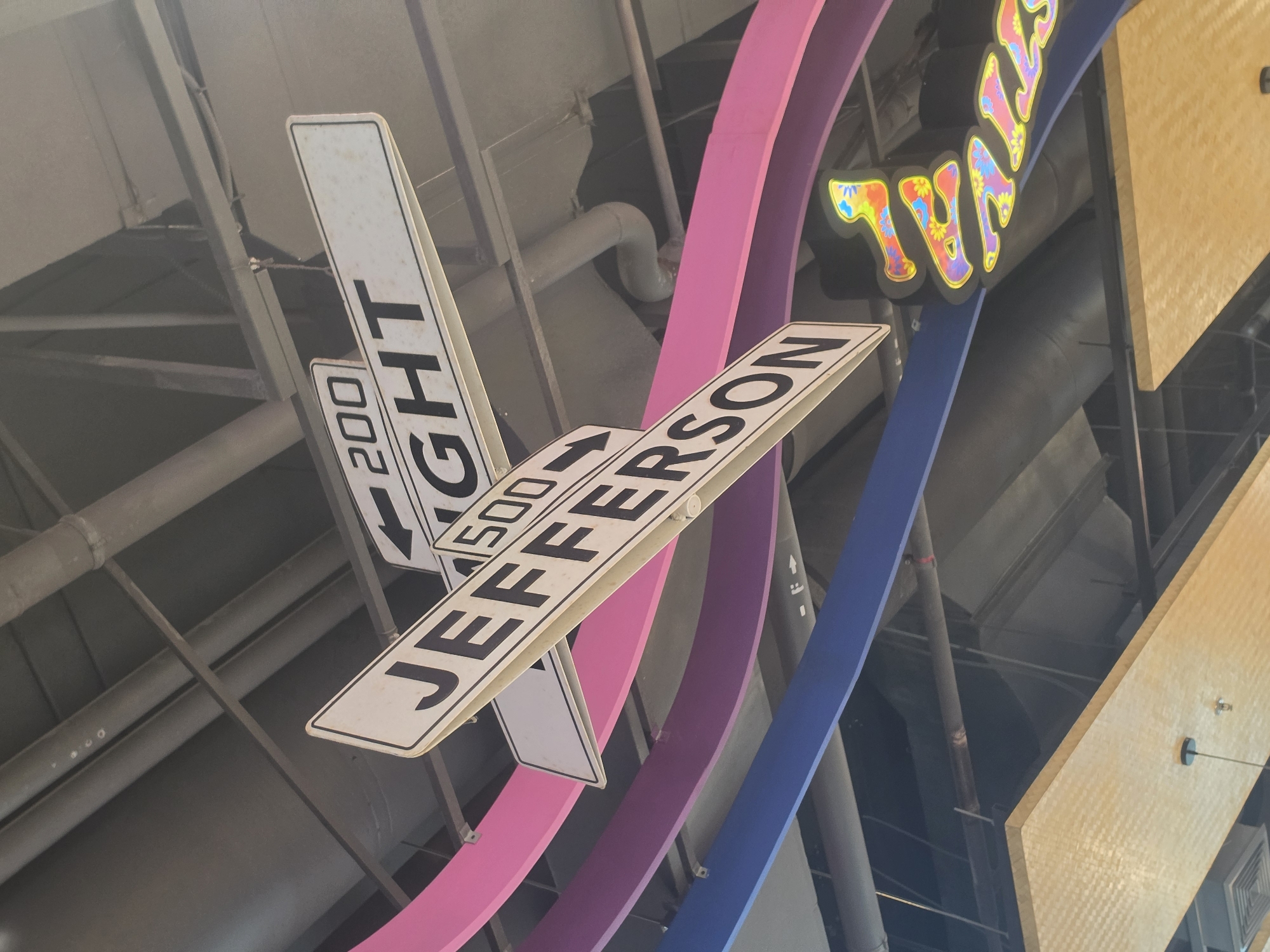 The image shows a cluster of street signs hanging from the ceiling inside a building. The signs read "1500 Jefferson" with an arrow and "200 Right". The signs are white with black lettering. Also visible are colorful decorative elements, including pink and blue curving shapes, and a portion of a sign with psychedelic lettering spelling "FESTIVAL". The ceiling appears to be industrial with exposed pipes and beams. No people are visible. The location in Pattaya City, Thailand cannot be confirmed or refuted solely from the image.