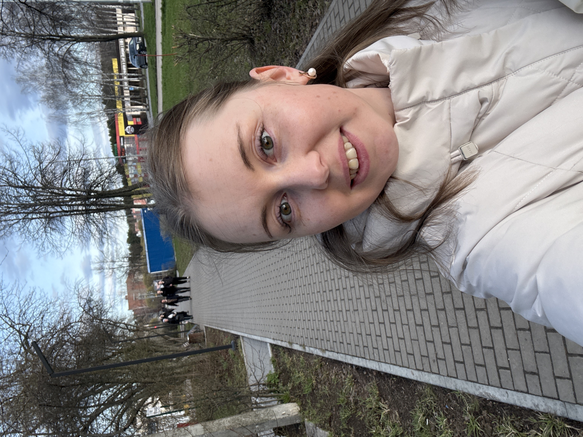 A woman is taking a selfie outdoors on a paved sidewalk. She is wearing a light-colored puffy jacket. In the background, there are trees with bare branches, a blue building, and a group of people walking away from the camera. A multi-colored building is in the distance and a car can be seen parked nearby. The location appears to be a city, but the specific city and area are not identifiable.