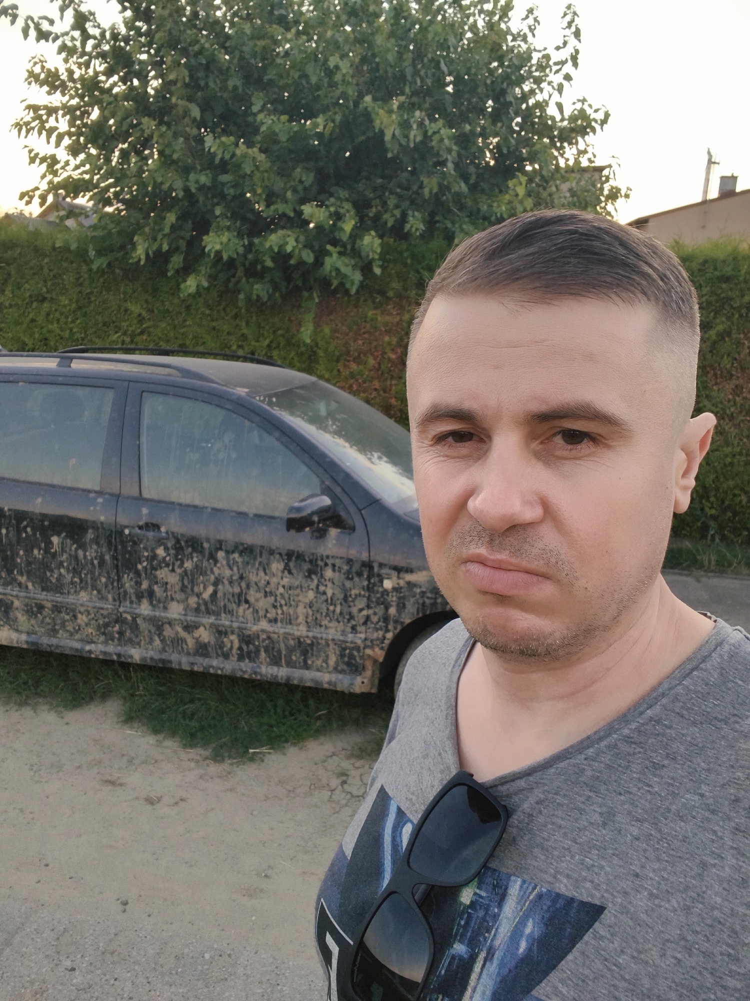 In the city of Tczew, Poland, a man stands in front of a dark-colored station wagon heavily covered in mud. The man is positioned in the foreground, facing the camera with a slightly downturned mouth and wearing a grey t-shirt with sunglasses clipped to the front. The station wagon, the primary object behind him, shows significant mud splatter across its body and windows, indicating recent travel through a muddy environment. Behind the car and man, a dense green hedge runs horizontally, and behind that, trees with lush foliage are visible against a light sky. The ground in front of the car appears to be a mix of dirt and gravel.