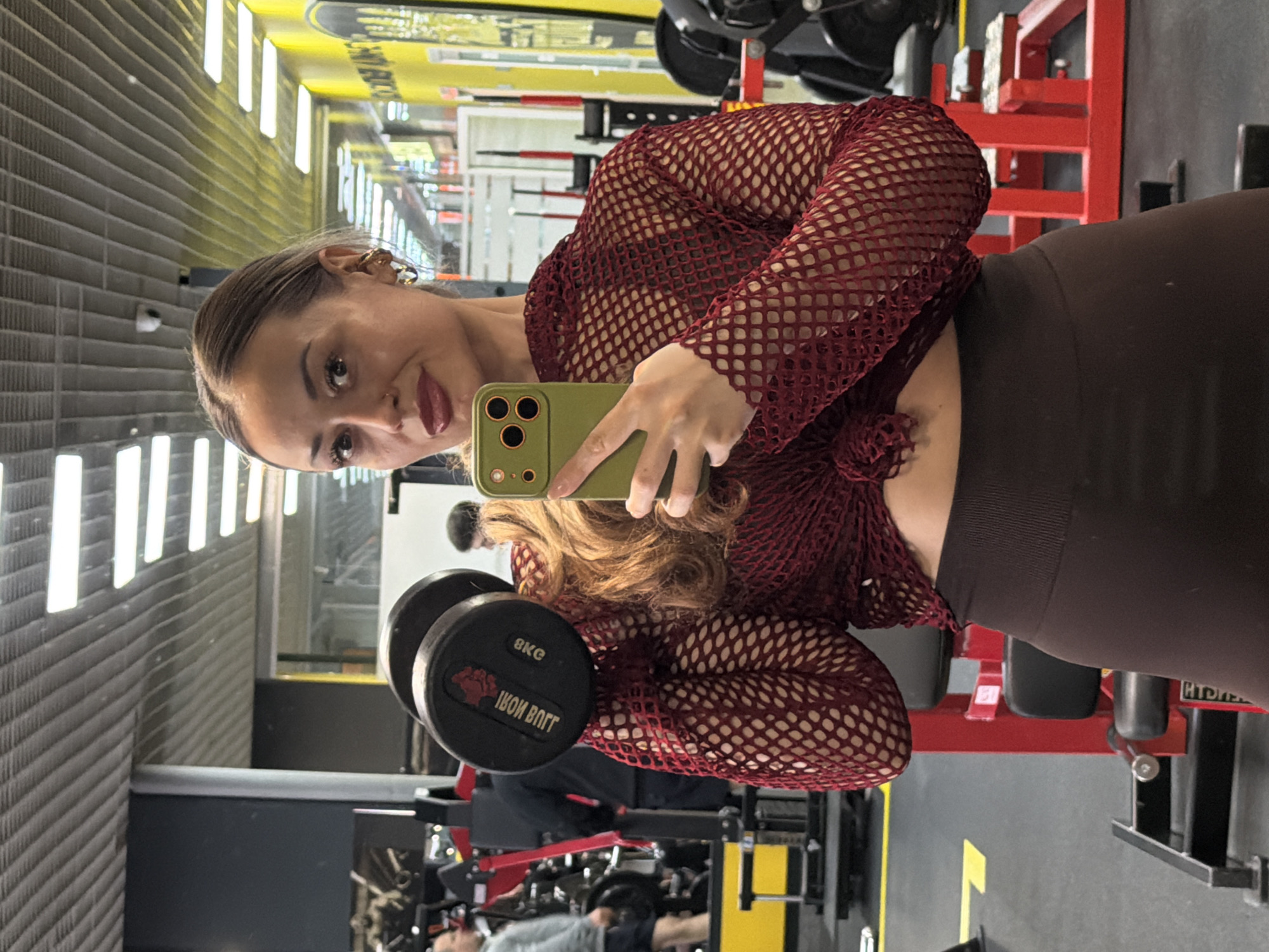 A woman stands in a gym, facing a mirror. She is holding a green phone in her left hand, taking a selfie. In her right hand, she holds a black 8kg dumbbell. She wears a maroon fishnet top and brown leggings. The gym contains various weight-training machines and equipment. Other people are visible in the background. The location is likely a fitness center in Krasnoyarsk, Russia.