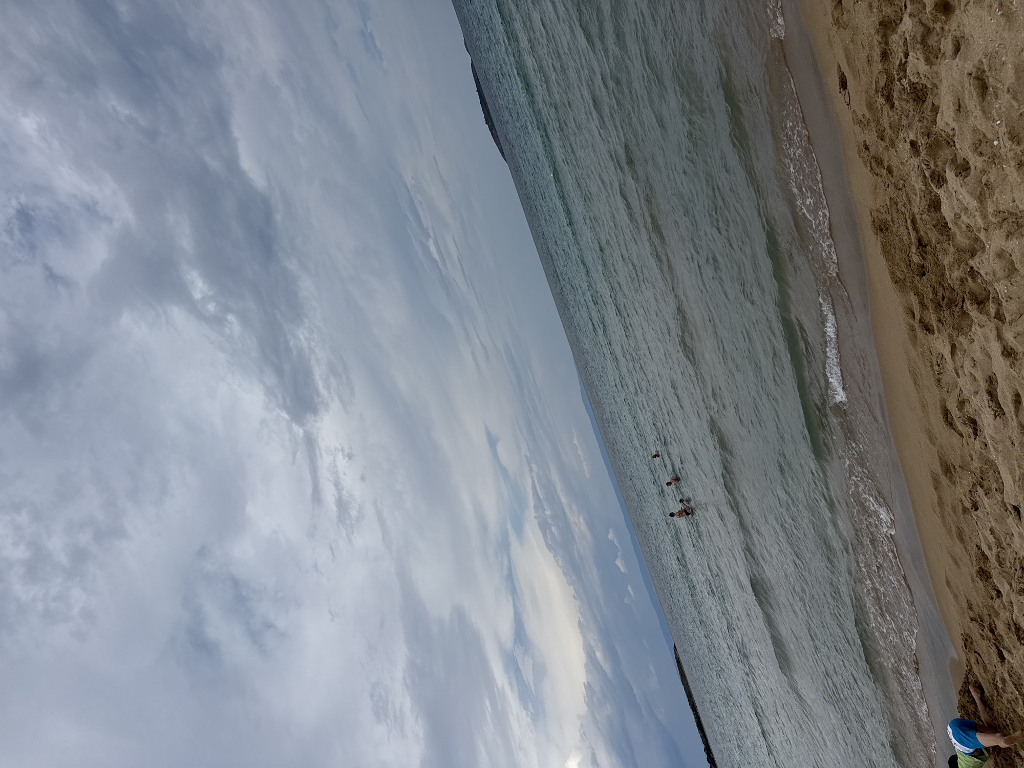 The image depicts a beach scene with a child in the foreground and several people in the water. The child, wearing blue and green clothing, is kneeling on the sand, appearing to play or dig. The sea is calm with gentle waves reaching the shore. In the distance, a few individuals are visible wading or swimming in the water. The sky is overcast with dramatic clouds, suggesting an approaching storm or a moody weather day. The location is the beach in Sozopol, Bulgaria, characterized by its sandy shore and the vast expanse of the Black Sea.