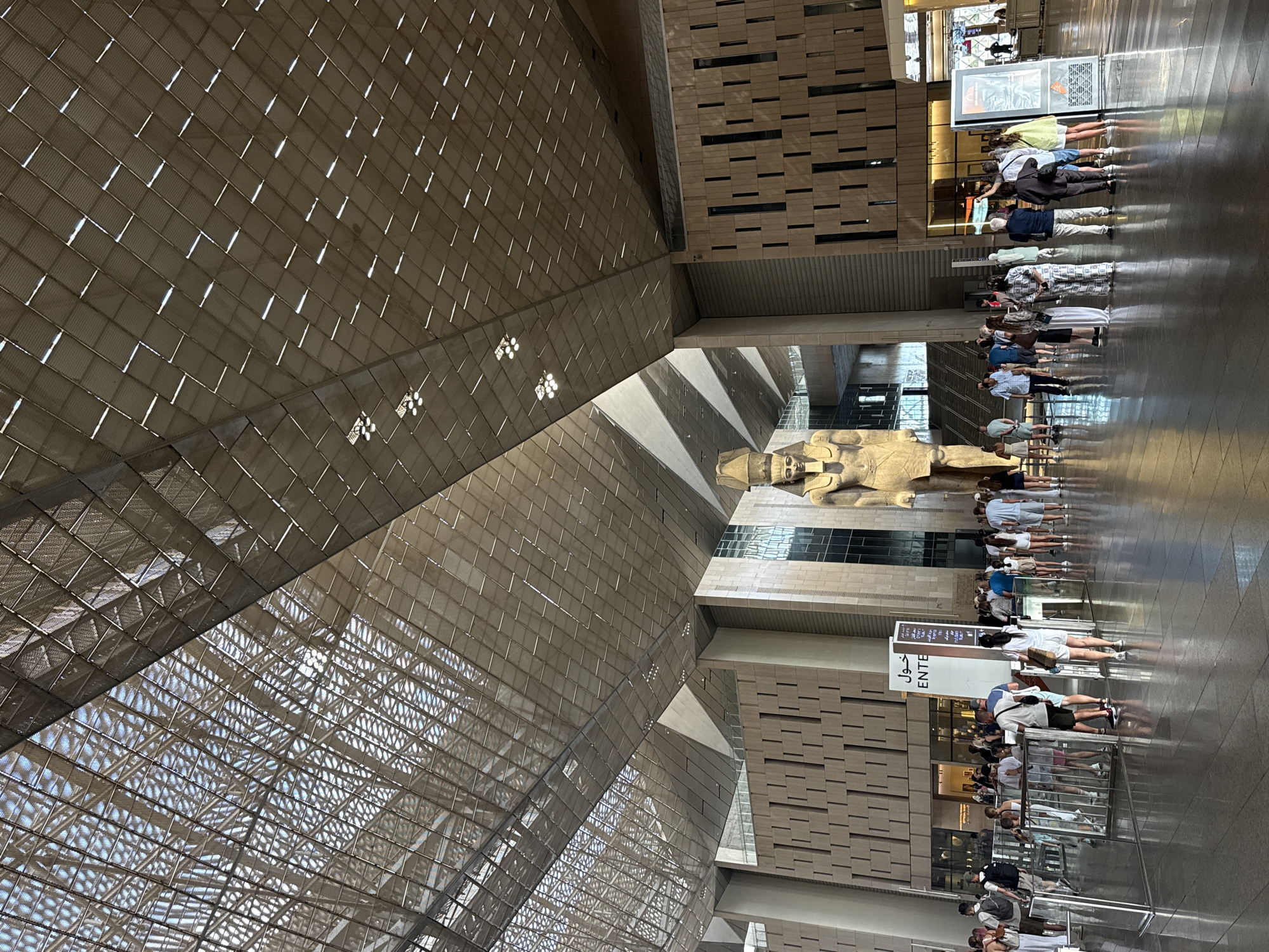 The image depicts the interior of a grand, modern museum or exhibition hall, likely the Grand Egyptian Museum, located in Kafr Nassar, Egypt. The architecture is characterized by a striking, geometric, sloped ceiling made of metal and translucent panels, allowing natural light to filter through. Large, imposing statues of ancient Egyptian pharaohs stand as central exhibits. In the foreground, a diverse crowd of tourists, dressed in casual summer attire, are gathered, appearing to be observing the exhibits or navigating the space. Some individuals are holding cameras or phones, suggesting they are documenting their visit. The floor is highly reflective, contributing to the spacious and luminous feel of the environment. Signage in both Arabic and English, including the word "ENTE" (likely for "entrance" or "enter"), indicates a public access area. The overall scene conveys a sense of awe and cultural exploration within a contemporary architectural setting.
