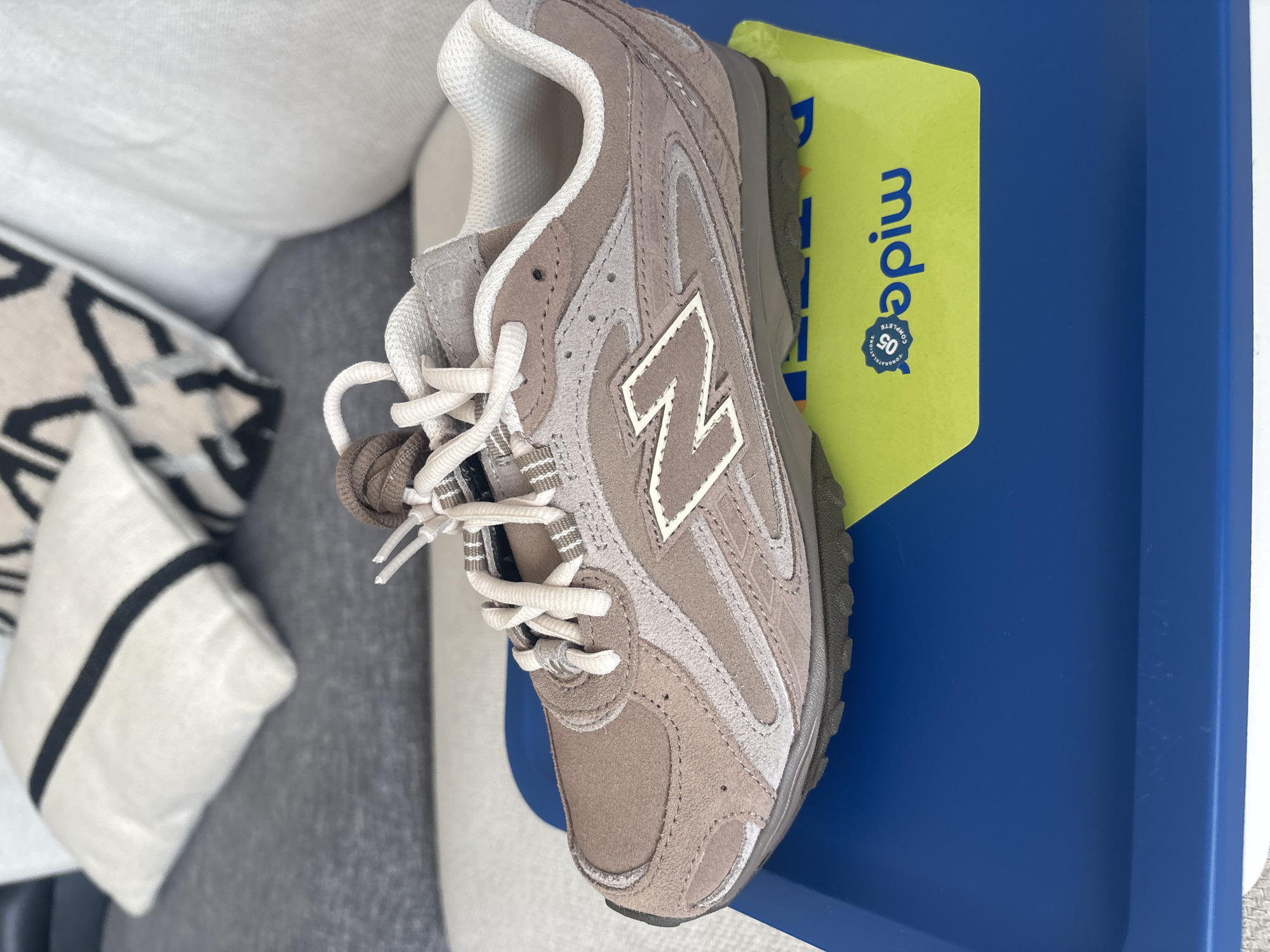 A single New Balance sneaker, primarily light brown with grey and white accents, rests on a bright blue tray. The shoe is placed on a yellow insert with blue lettering that reads "05 COMPLETE". In the background, out of focus, are two decorative pillows on a grey sofa. One pillow is white with bold black stripes, and the other is solid white. The scene suggests a domestic setting, potentially in Dubai, United Arab Emirates.