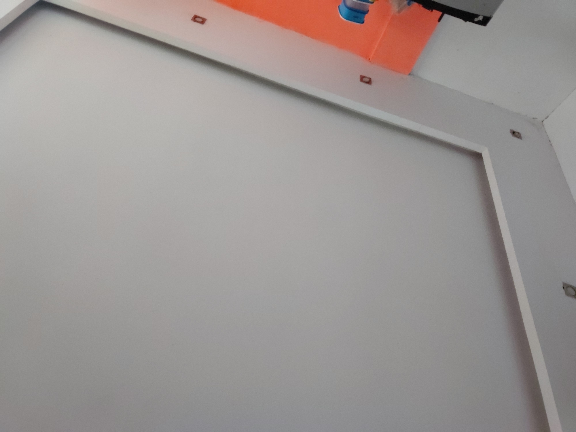 The image captures an interior architectural detail, showcasing a light gray wall framed by white decorative molding. This molding creates a large rectangular outline on the wall. Above the white trim, a vibrant orange wall or ceiling section is visible, suggesting a distinct color palette in the room.
Several small, square, recessed light fixtures, appearing to be unfinished or without their covers, are embedded into both the white molding and the orange surface. In the upper right portion of the image, a white ceiling is visible, along with a dark, possibly black, structure or piece of equipment hanging from it. A blue and white object, perhaps a container, rests on the orange surface in this area.
The perspective is angled upwards, emphasizing the junction between the walls and ceiling. No people are present, and no specific activity is occurring, presenting a static view of an interior space that might be under construction or renovation. There is no visible text within the image. Based on the provided context, the location is Santa Ana, Venezuela.