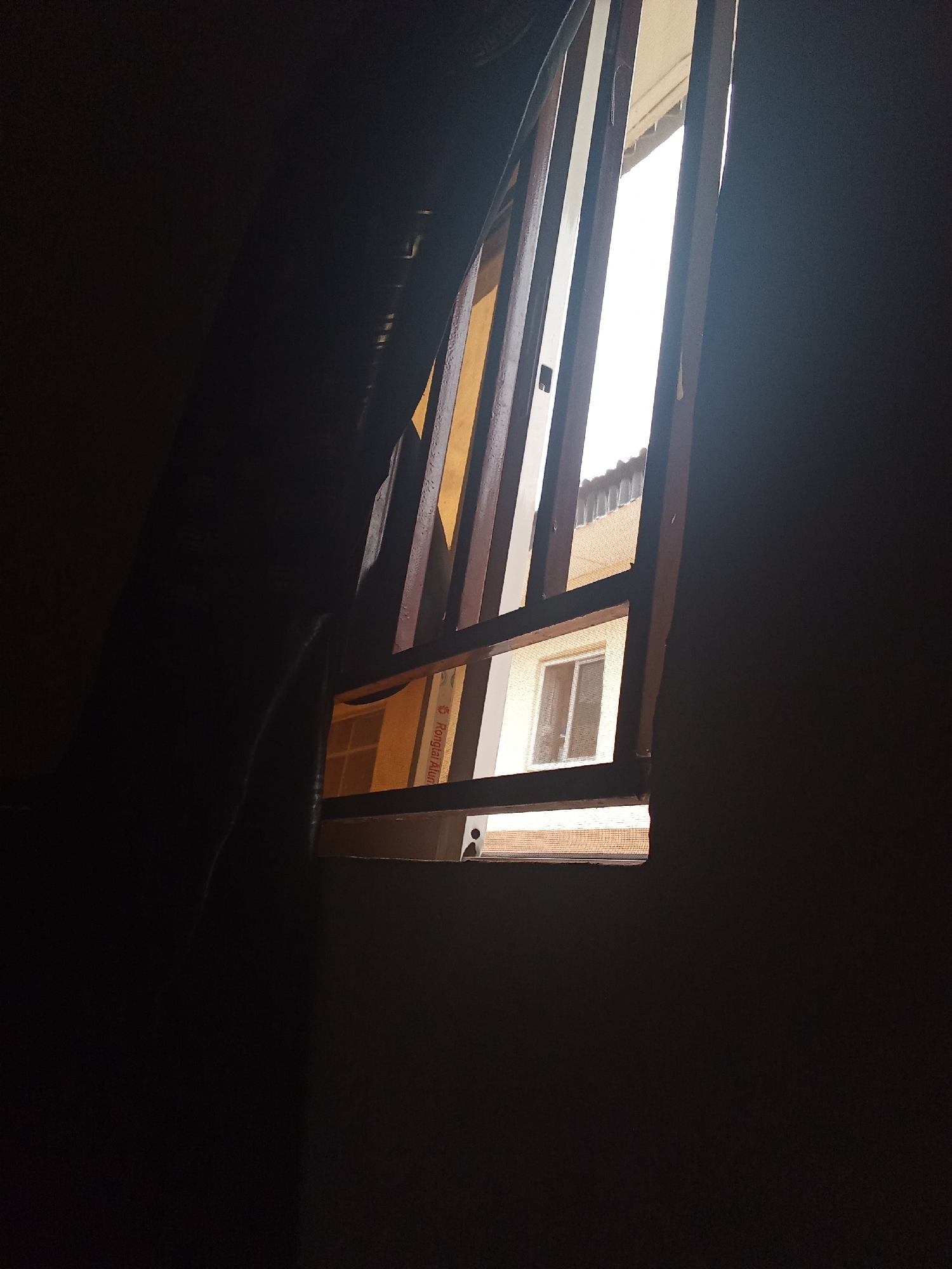 The image shows a view through a window with bars. The window is partially open, allowing bright light to stream in from the outside. In the foreground, a dark curtain or drape hangs on the left side of the frame, obscuring much of the interior space. The window itself has a wooden frame and metal bars, suggesting it might be a security window.
Through the open section of the window, the exterior scene is visible. There's another building with windows and a tiled roof. The bright light suggests it is daytime, possibly sunny. The angle of the light and the contrast between the bright exterior and the dark interior create a dramatic effect. The scene appears to be indoors, looking out.
There is some text visible on a piece of paper or card in the lower right section of the window, behind the bars. The text is in a dark color on a light background and appears to be in a foreign script, with some letters resembling "Rongiar Alun". The overall mood is somewhat mysterious due to the limited visibility and the stark contrast of light and shadow.