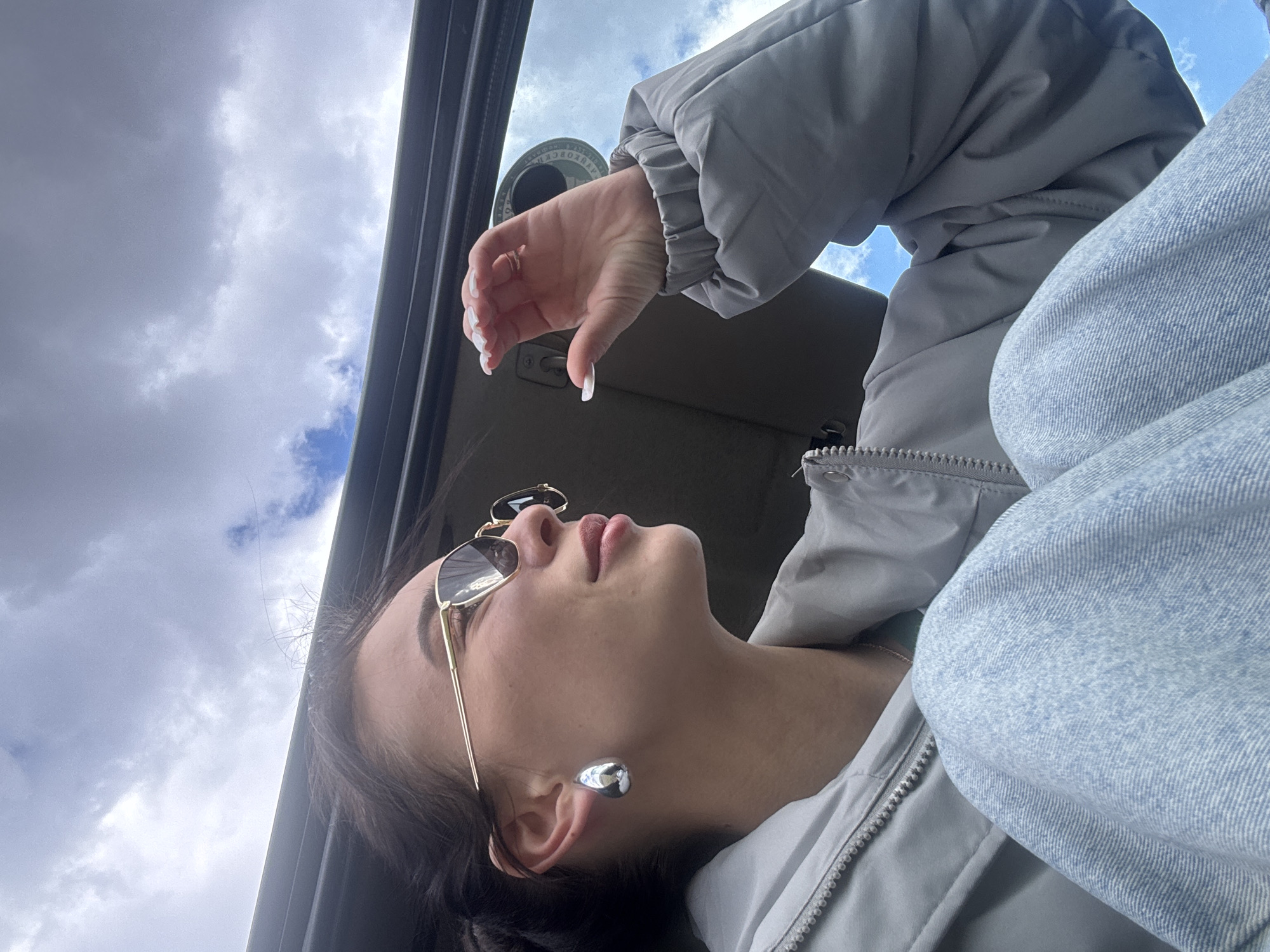 An overhead, slightly angled photo captures a person, likely female, posing inside a vehicle while looking upwards towards a cloudy sky. The individual is positioned on their back or reclined, with their head tilted up and to the right, showcasing their left profile. They wear stylish gold-framed, dark-lensed sunglasses and a light gray puffer jacket with a visible zipper. Underneath the puffer jacket, a light blue denim garment is visible on their arm/shoulder. A prominent, rounded, silver-toned earring adorns their left ear. Their hand, visible in the upper right, has long, light-colored nails and a small ring on one finger.
The background consists primarily of an overcast or partially cloudy sky, with some patches of brighter blue peeking through. The frame of a car window or sunroof is visible along the right side of the image, suggesting the person is looking out from a vehicle. Above the person's hand, a circular object with Cyrillic text is partially visible, appearing to read "НОВГОРОДСК" (Novgorodsk), a detail that aligns with the given location context of Фоки, Russia. The overall lighting suggests daytime. The person's expression, though partially obscured by sunglasses, appears neutral or contemplative as they gaze upwards.
