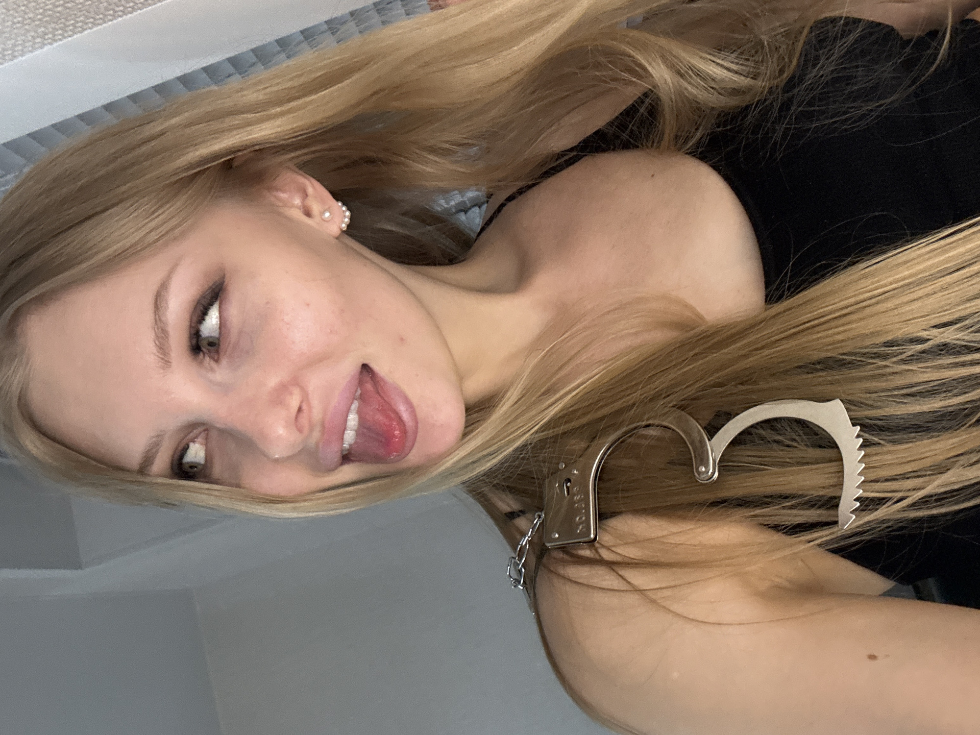 A light-skinned young woman with blonde hair and light eyes is shown in a close-up shot. She is wearing a black top and has what appears to be a handcuff-shaped hair clip in her hair. Her tongue is sticking out. The background shows a gray wall and a light blue and white striped surface. There is no clear indication in the image that the location is in the city of Perm, Russia.