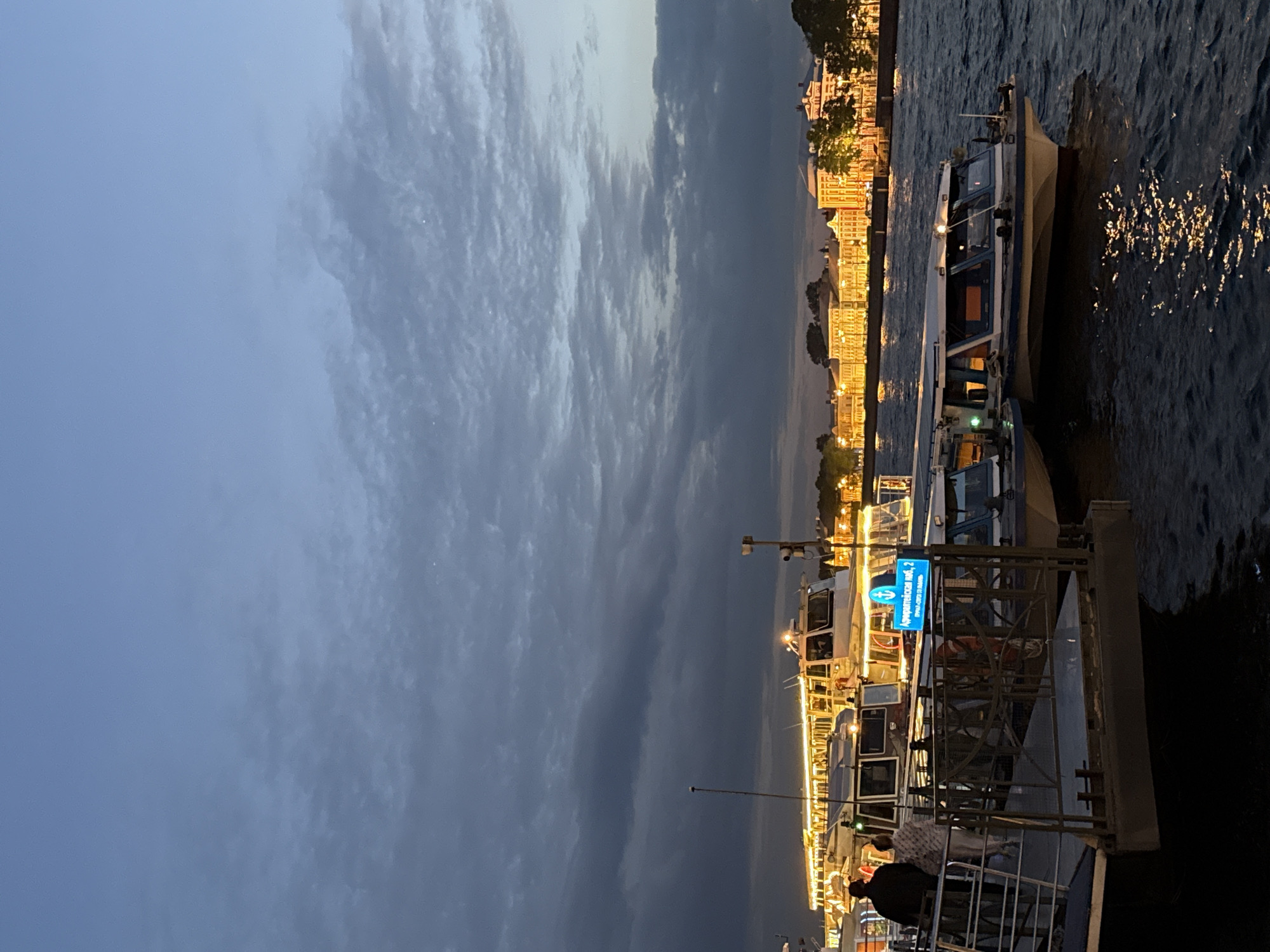 This image depicts an evening or twilight scene along a river or canal in Saint Petersburg, Russia. The sky, covering the upper portion of the frame, is predominantly overcast with varying shades of dark and light grey clouds, indicating either dusk or early night.
On the right side of the image, several passenger boats are docked, with their interiors and decks brightly lit, casting reflections on the dark water. The water itself shows shimmering streaks of light from these illuminations and potentially from distant sources. Across the river, the opposite bank features a row of buildings with numerous illuminated windows and facades, glowing warmly against the darkening landscape. The architectural style of these buildings suggests a European urban setting.
Two individuals are partially visible in the lower right foreground, appearing to be on or near one of the boats or a docking platform.
A notable detail is a blue sign visible on one of the boats or the dock structure. The text, written in Cyrillic, clearly states "Адмиралтейская наб., 2" (Admiralteyskaya Embankment, 2), with "Речные прогулки" (River walks/cruises) below it. This text strongly identifies the location as Admiralteyskaya Embankment in Saint Petersburg, a known area for river tours. The presence of multiple passenger boats further suggests an activity of river cruises.
The overall mood is tranquil, characterized by the contrast between the artificial light from the boats and buildings and the natural fading light of the cloudy sky.