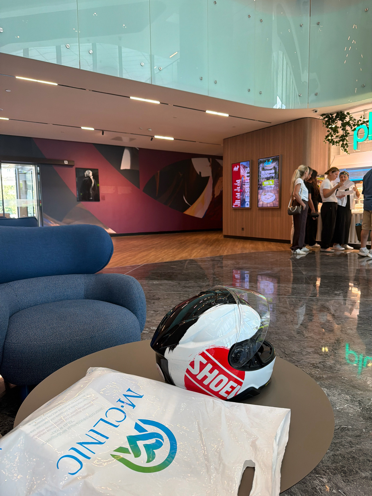 The image depicts a modern interior space, likely a shopping mall or a public lounge area, situated in Ataşehir, Turkey. In the foreground, a round, taupe-colored table holds a white plastic bag with blue and green lettering that reads "M.CLINIC" and a graphic logo, alongside a white and black motorcycle helmet with a prominent red "SHOEI" logo. To the left, a plush blue armchair is partially visible.
The background reveals a polished, dark marble floor with reflections. Further back, a wall features a large, abstract mural in shades of maroon, gray, and white, with a framed portrait of a person in black and white attire. Beyond this, a set of illuminated digital display boards showcase advertisements, one reading "DESSERTS" and another with Turkish text and a price.
To the right, several individuals are standing, appearing to interact with a counter or another person out of frame. The architecture includes a curved wooden wall and a high ceiling with a reflective, green-tinted glass enclosure, illuminated by linear light fixtures. A hanging plant arrangement is visible near the ceiling. The overall impression is a clean, contemporary, and well-lit public space.