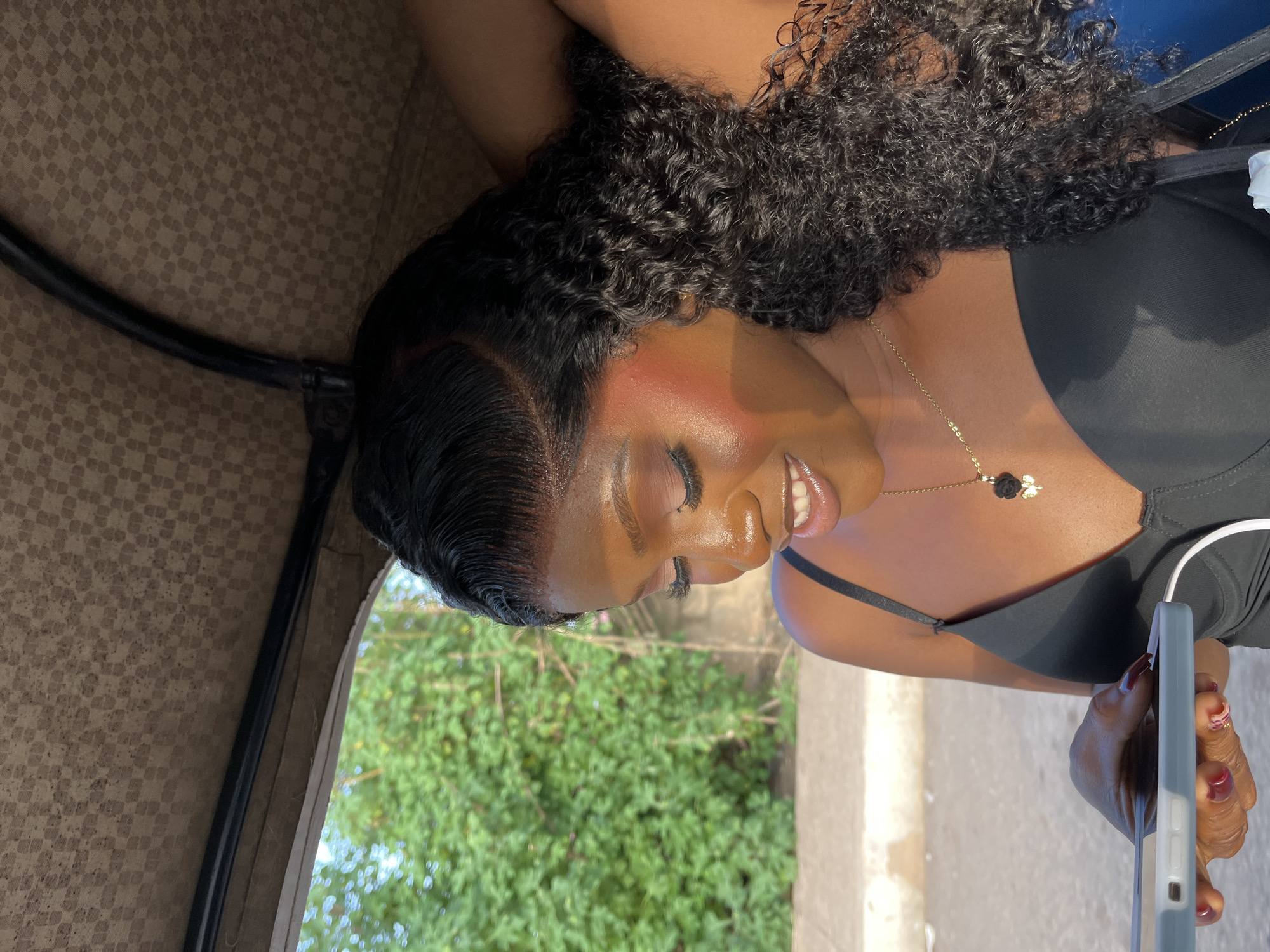 A dark-skinned female, likely a young adult, is depicted in a close-up, smiling while looking down at a smartphone held in her left hand, in the city of Gura Top, Nigeria. She has long, dark, curly hair with a defined side part and a visible lace front. Her face exhibits makeup, including prominent blush on her cheekbones, eyeshadow, and long eyelashes. She wears a black bra or camisole with thin straps and a thin gold necklace with a black floral pendant and a small gold charm. Her fingernails are painted dark red. The smartphone has a light grey and white/light blue two-toned case, with a white earbud wire connected. She is situated inside a vehicle, indicated by a beige fabric roof with a subtle checkered pattern and a black metal support bar visible overhead. The outdoor background features lush green foliage and trees, suggesting daylight conditions.