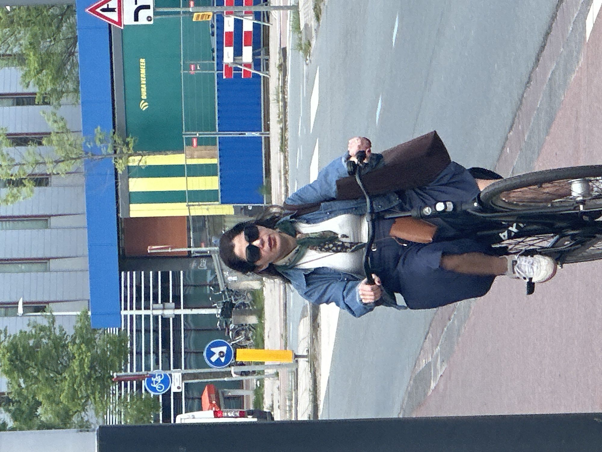 The image shows a woman riding a bicycle on a city street. She is wearing sunglasses, a denim jacket, and a skirt. She is also carrying a brown bag. The background includes buildings, street signs, and other bicycles. The location appears to be Amsterdam, Netherlands, based on the architecture and street signs. There is a 'Dura Vermeer' sign visible in the background. The woman is actively cycling. The street signs indicate bike lanes.