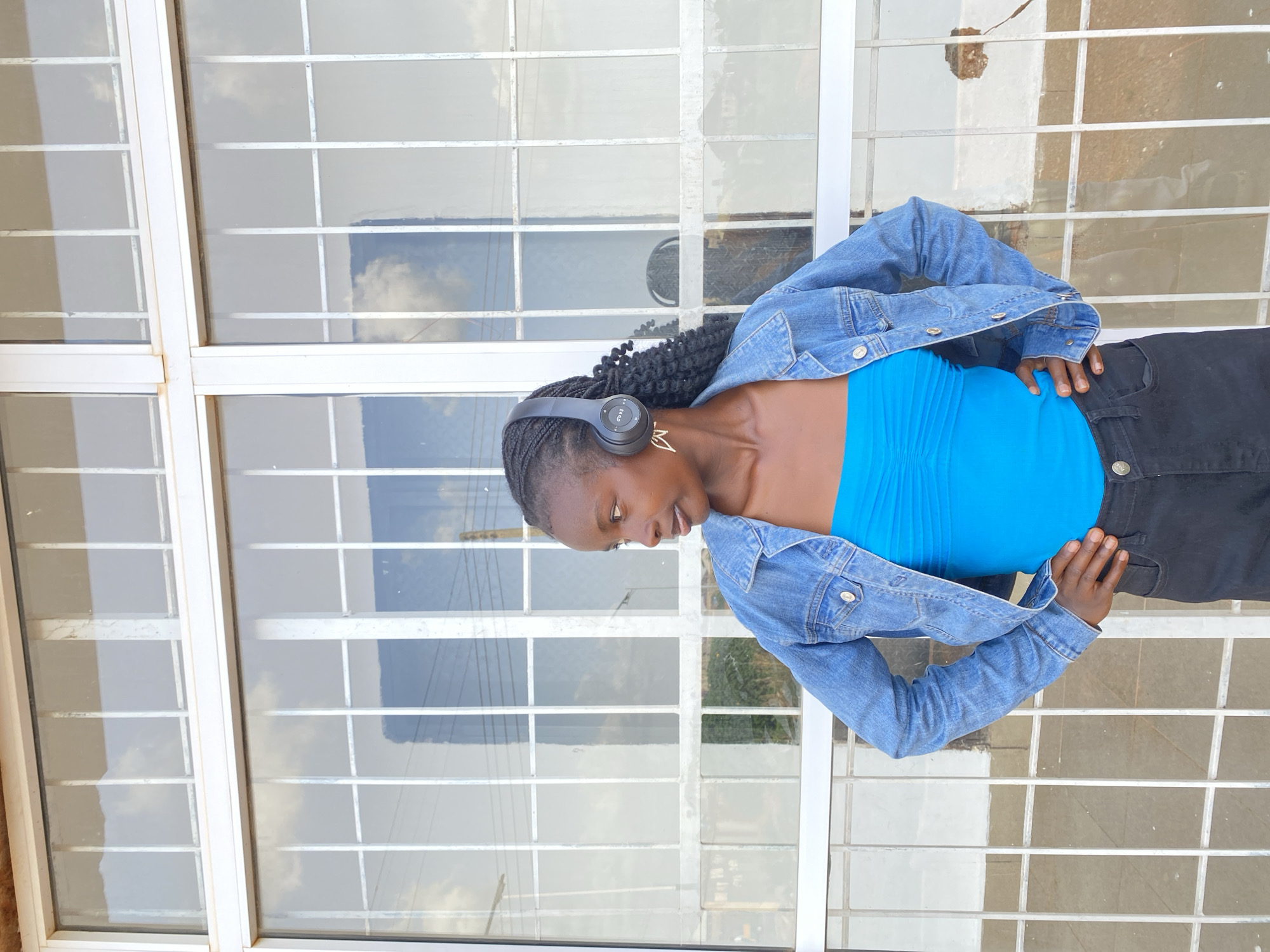 A young Black woman stands in front of a window with white bars, wearing a blue tube top, a denim jacket, and black pants. She has headphones on her ears and is looking to her left with her hands on her hips. The window reflects the sky and some buildings, and the background appears to be outdoors in an urban setting.