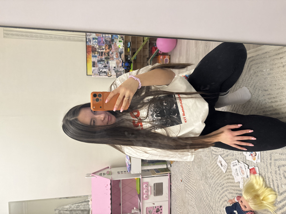 A young woman is reflected in a mirror. She is kneeling and facing the mirror, holding up an orange phone. She wears a white t-shirt with a graphic, black leggings, and one white sock. Her long dark hair hangs down. Behind her, visible in the reflection, is a room with a pink toy kitchen, playing cards scattered on a carpet, a blonde doll, and shelves with toys. A collage is visible on the wall. She has a lilac wristband.