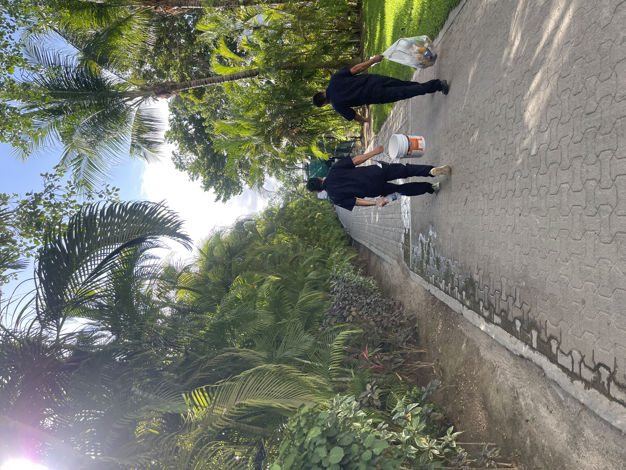 Two people in dark uniforms walk along a paved walkway in Playa del Carmen, Mexico. One person carries a clear plastic bag filled with trash. The other person carries a white bucket in their left hand and a water bottle in their right hand. The walkway is bordered by a stone wall and lush green vegetation. Palm trees are visible in the background. The weather appears to be sunny.