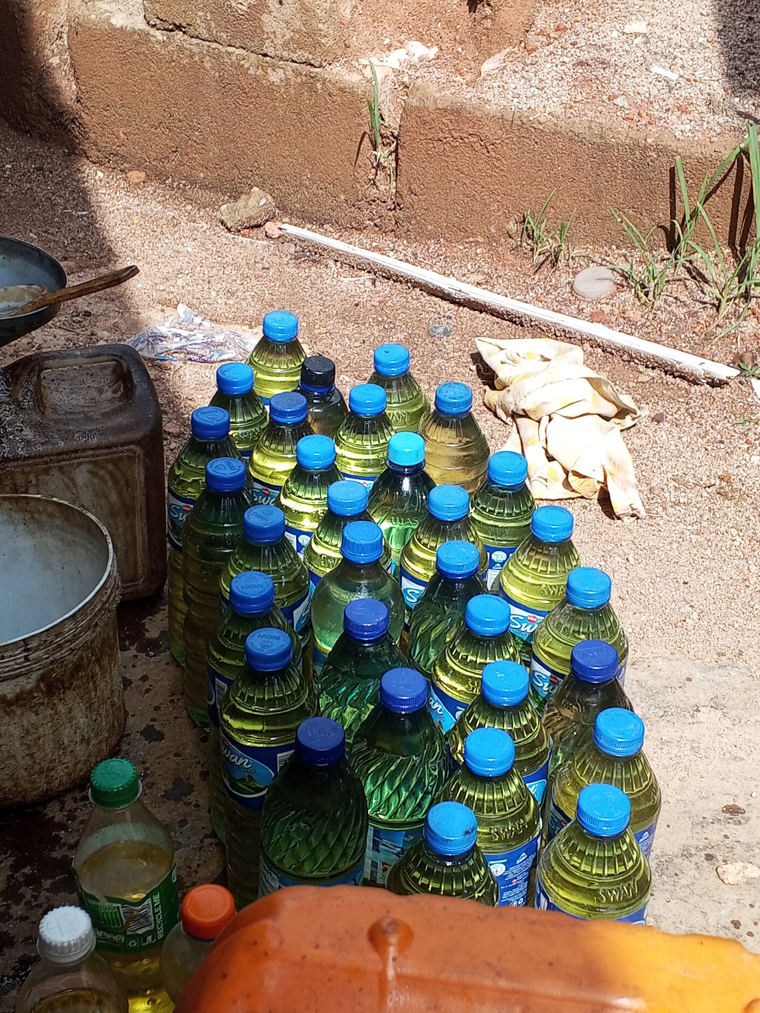 The image shows a collection of approximately 25 plastic bottles filled with a yellow liquid, likely cooking oil. Most of the bottles are branded "SWAN" and have blue caps, while a few near the bottom have green, white, and orange caps. The bottles are arranged closely together on a gravelly surface. A dirty, white cloth is lying next to the bottles. To the left are a couple of used containers. A rusty pan with a wooden spatula sitting in it is in the upper left. The backdrop includes a dirt wall with some sparse green vegetation at the top.
The location is Gwong, Nigeria. The scene suggests a possible roadside or informal market setup for selling cooking oil. The environment is dusty and somewhat unkempt. No people are visible in the image.