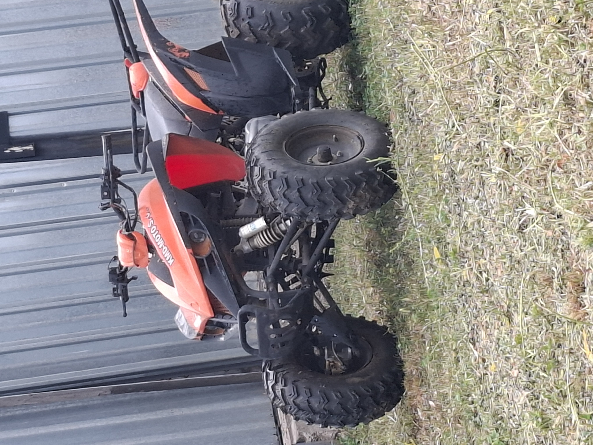 An orange and black ATV is parked on a patch of grass in front of a corrugated metal wall. The ATV is facing left, with its front wheels turned slightly to the right. The ATV has a black handlebar and seat, with orange accents on the front and rear fenders. The ATV has large, knobby tires. The corrugated metal wall is grey and appears to be part of a building or fence. The grass is green and appears to be slightly overgrown. The scene is outdoors, and the lighting suggests it is daytime. There is no indication of the specific location in Almaty, Kazakhstan.