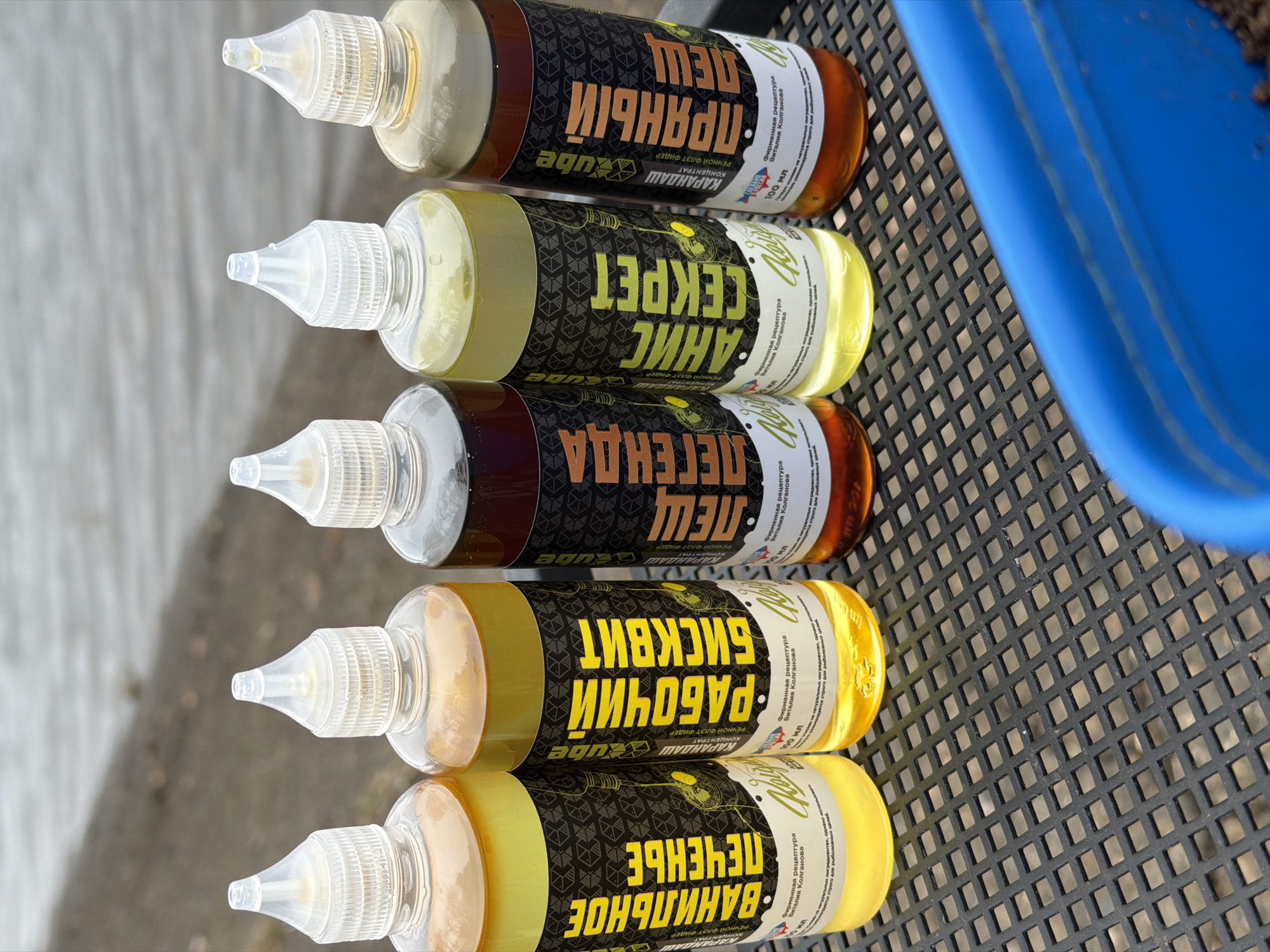 The image shows five bottles of fishing attractants stacked on a black, latticed surface. The bottles have clear caps and labels in Russian. The labels are for: "Vanilla cookies," "Working biscuit," "Legend, Bream," "Anise secret," and "Spiced, Bream." A blue object is partially visible to the right of the bottles. The location is Chaikovsky, Russia.