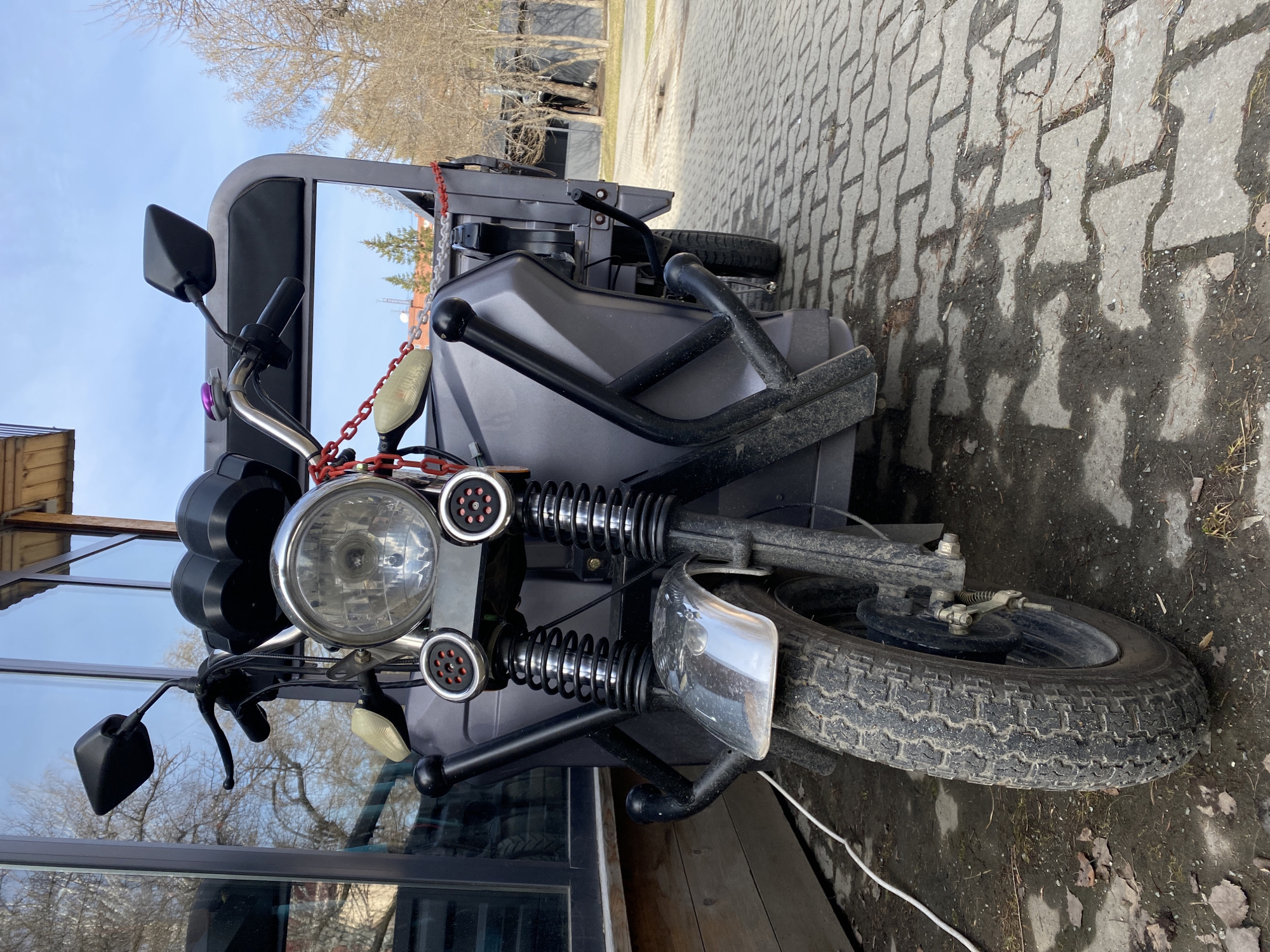 The image depicts a gray tricycle-like vehicle with a passenger area and a prominent headlight. It is equipped with spring-loaded shock absorbers, rearview mirrors, and a red chain. The vehicle is parked on a paved surface of rectangular bricks. The surrounding environment includes buildings with mirrored windows reflecting trees and a clear sky. The presence of birch trees suggests a location in or around Yekaterinburg, Russia.