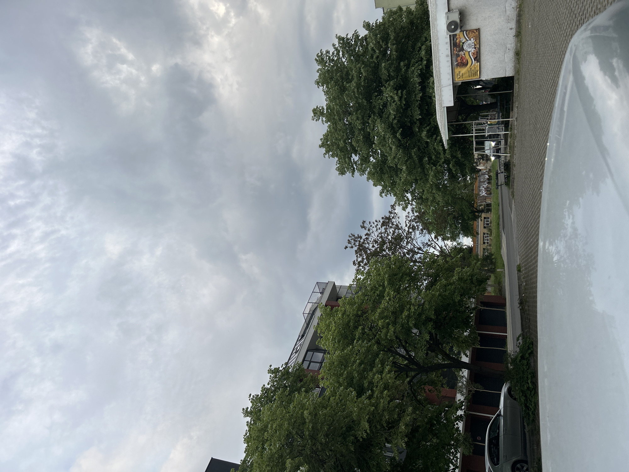 The image shows a street view in what is purported to be Burgas, Bulgaria. Overcast skies dominate the upper half. On the right, a partially visible white vehicle fills the foreground. A building with a fast food advertisement is also on the right, with tables and chairs outside. In the middle, trees line the street, partially obscuring a modern building with balconies. A gray car is parked beside the building.