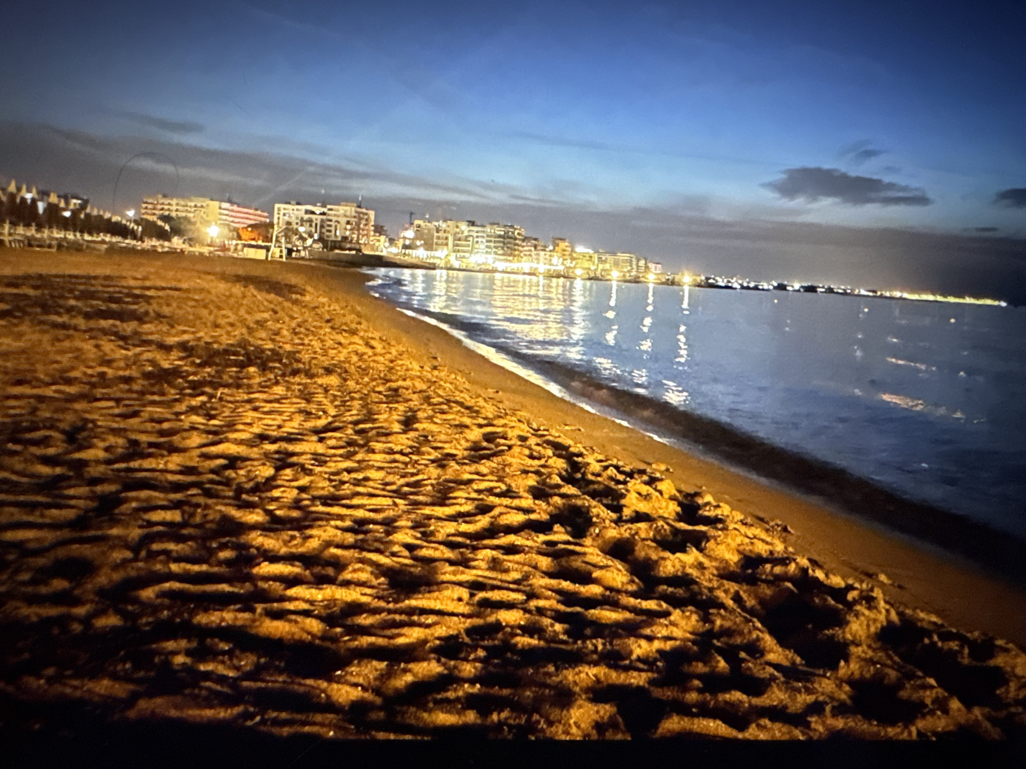 The image depicts a beach at twilight, with the lights of a coastal city illuminating the background. The foreground is dominated by the textured surface of wet sand, reflecting the artificial lights from the city. Gentle waves lap at the shore, their surfaces shimmering with reflected light. The sky is a deep blue, transitioning from the brighter horizon to a darker zenith, with a few wispy clouds visible. The city skyline, composed of multi-story buildings with numerous illuminated windows, stretches along the coast. The scene conveys a sense of quiet evening ambiance by the sea. The image does not depict the city of Porrentruy, Switzerland, as Porrentruy is an inland town and not located on a coast.