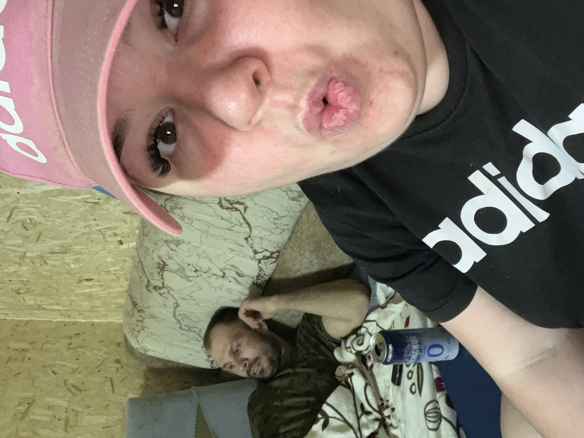 A selfie photograph depicts a woman in close proximity to the camera, wearing a pink baseball cap and a black Adidas t-shirt, puckering her lips. Behind her, a man is lying down on a bed or couch, covered with a blanket. Beside him is an open can of "Anchor" brand beer. The setting appears to be indoors with light-colored wood paneling. The image was taken in the city of Chaikovsky, Russia.