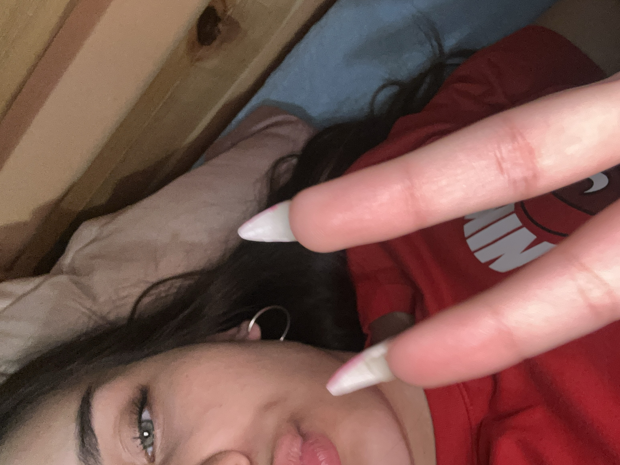 The image shows a close-up of a young person's face and hands. The person has dark hair, green eyes, and is wearing a red t-shirt. They are making a peace sign with their right hand, with long, pointed, light-colored fingernails. Their left hand is also visible, also with similar fingernails. The background appears to be a bed with a pillow and a wooden headboard. There is no indication of the location being the city of Shumen, Bulgaria.
