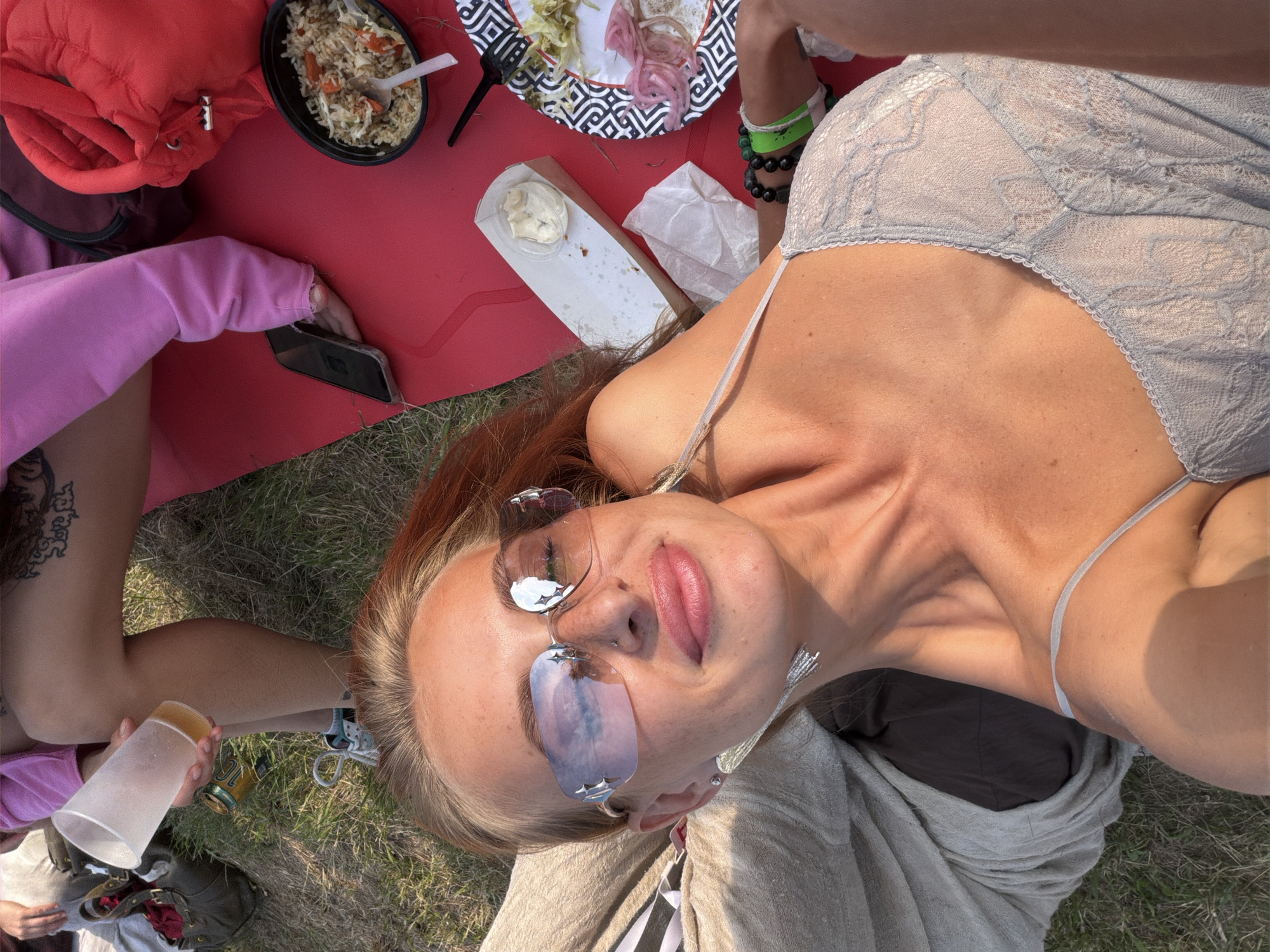 The image shows a group of people having a picnic outdoors. The scene is set on a grassy area, likely a park or field, with a red picnic blanket spread out. Food items are visible, including a bowl of rice and vegetables, a plate with food remnants, and a container of dip. One person, with reddish-blonde hair, is wearing unique star-shaped sunglasses and a lacy top. They are looking up at the camera, with their mouth slightly open. Another person, wearing a pink hoodie, is holding a smartphone and a translucent cup. There are also some cans and bags visible. The lighting suggests it is daytime, likely late afternoon due to the warm glow and shadows. The overall mood appears relaxed and casual, indicative of a leisurely outdoor gathering. The location context is Дрокино, Russia. There is no visible text in the image.